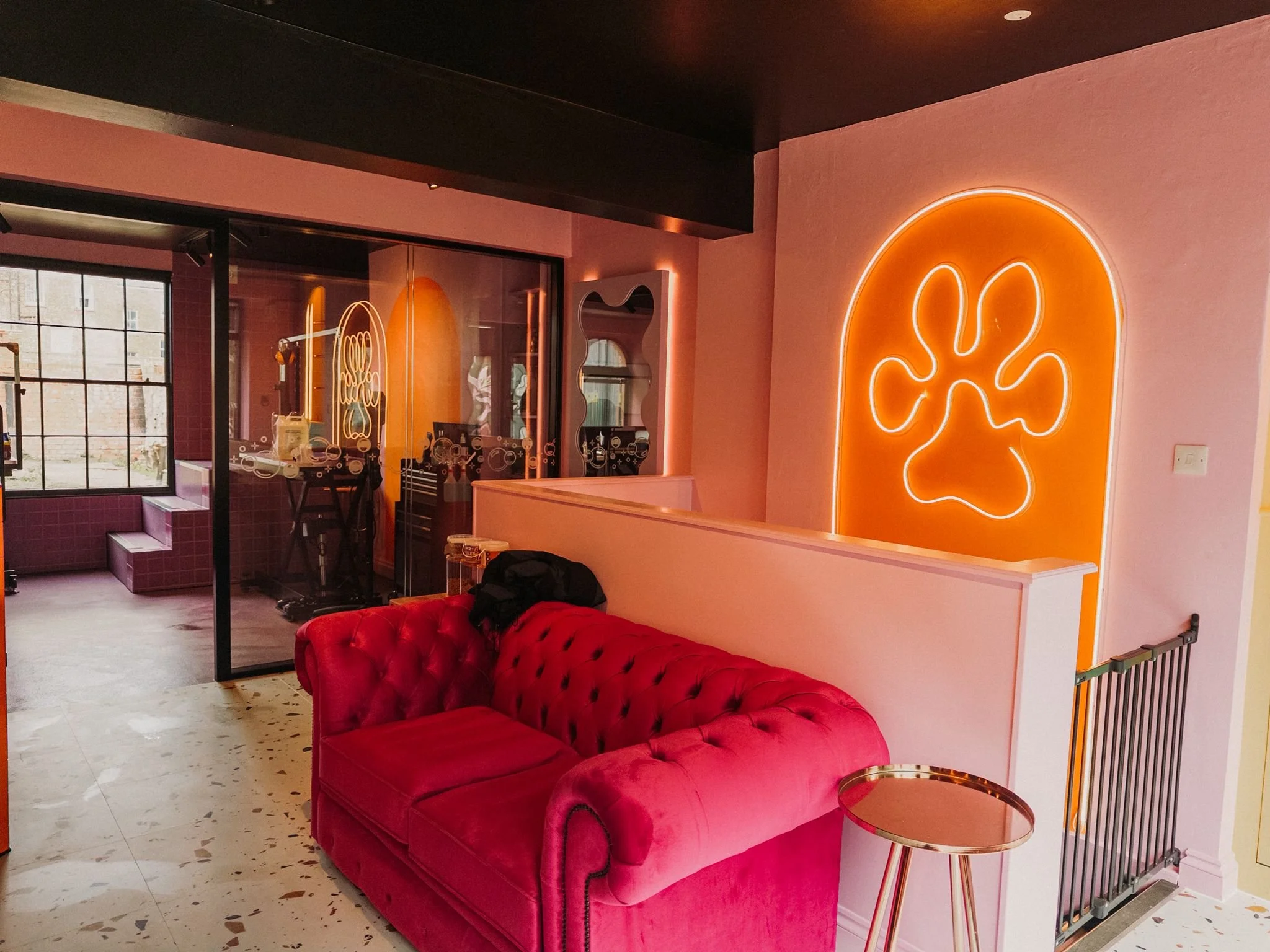 Interior of a trendy cafe with a neon orange plant-shaped sign on a pink wall, a red velvet tufted sofa, a small round table with a gold rim, and large windows showing an exterior brick building.