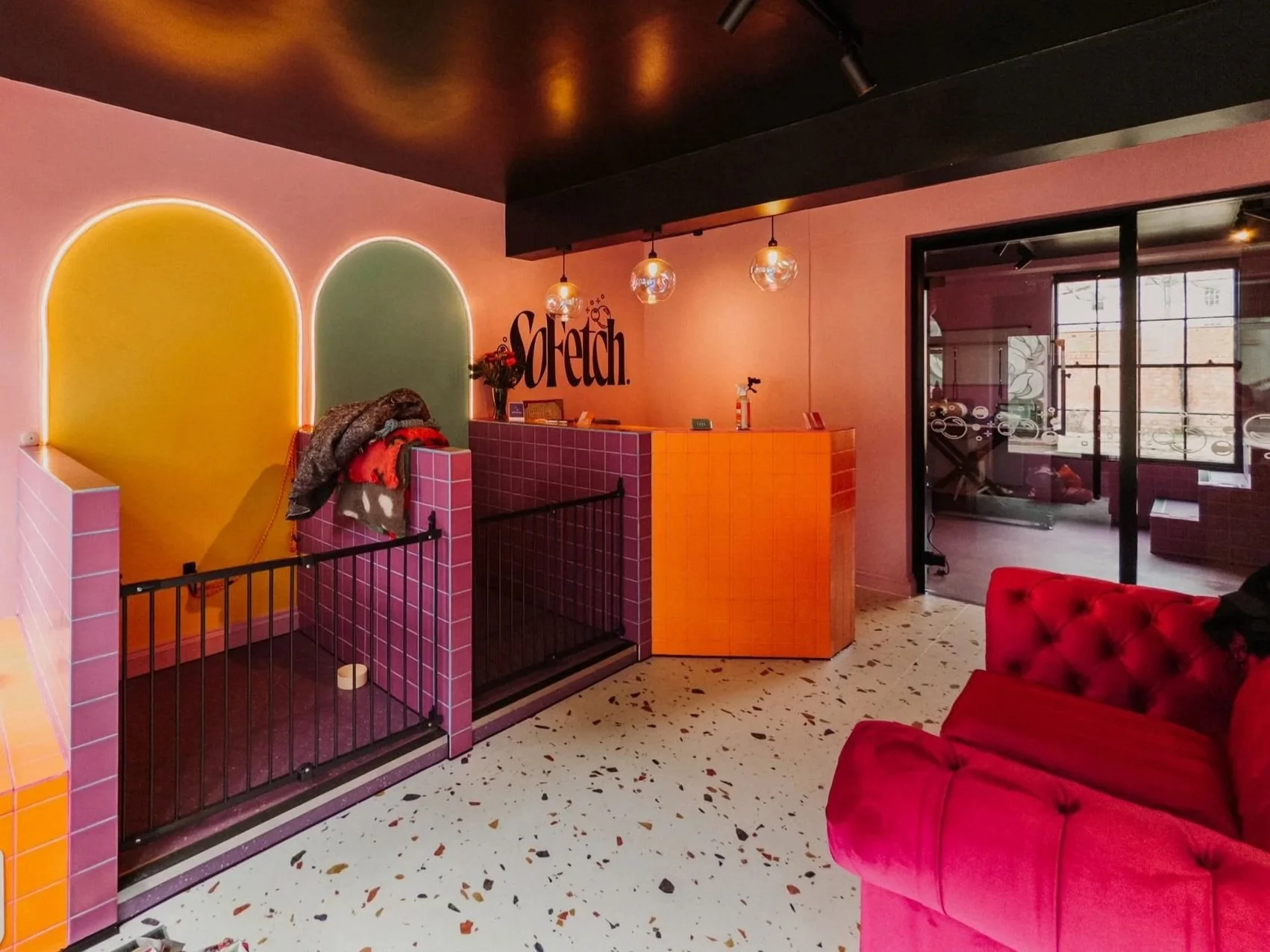 A brightly lit, colorful interior space with pink and orange tiled walls, a pink tiled reception desk, and a red tufted sofa. There are two arched wall niches painted yellow and green with backlighting, and glass doors lead to a gym area with exercise equipment visible through the glass. Hanging globe lights are above the reception area.