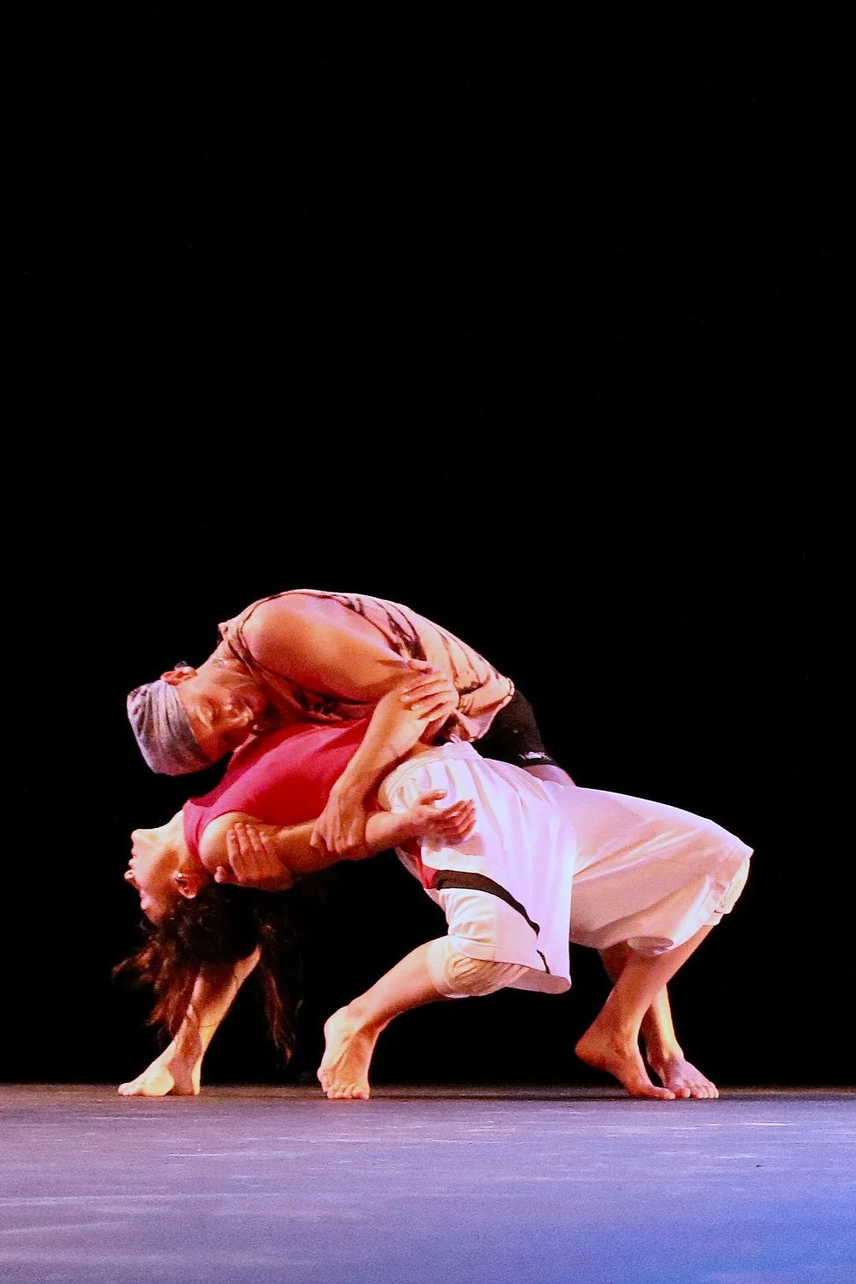 A man and a woman are dancing on a dark stage, intertwined in a passionate pose, with one wearing a pink top and gray head covering, the other in a white skirt and black top.