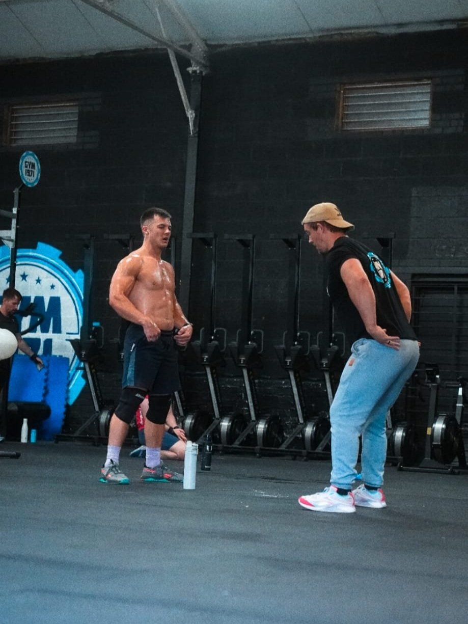 A shirtless man in workout clothes talking to a man in a black shirt and gray sweatpants inside a gym. The man in sweatpants is bowing slightly with hands on his hips. The gym has black walls, equipment, and a blue and white logo in the background.