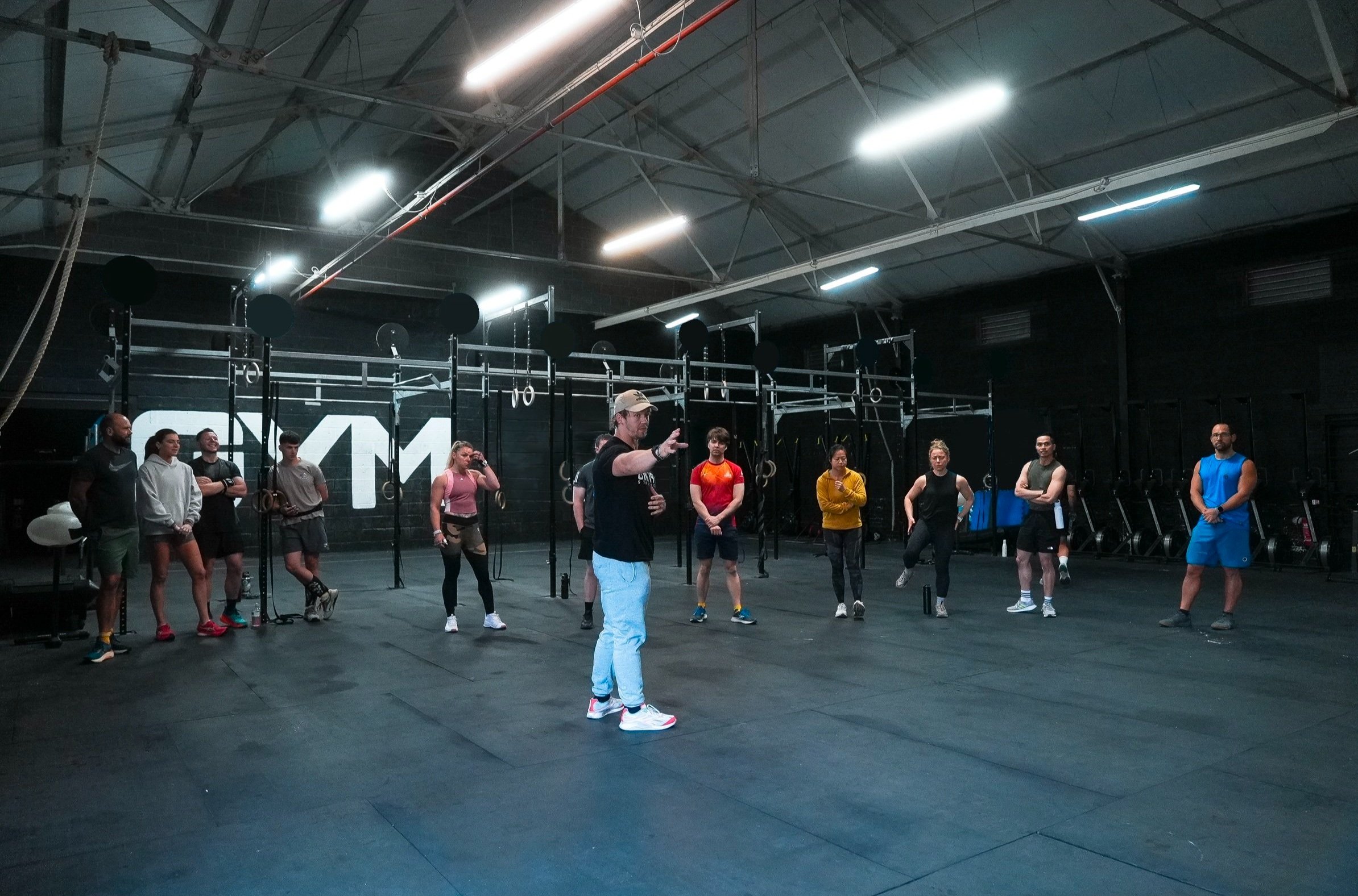 A group fitness class is taking place in a gym with a coach leading an exercise. The gym has black walls, gym equipment, and a large logo that reads 'GYM 1971' on the walls.