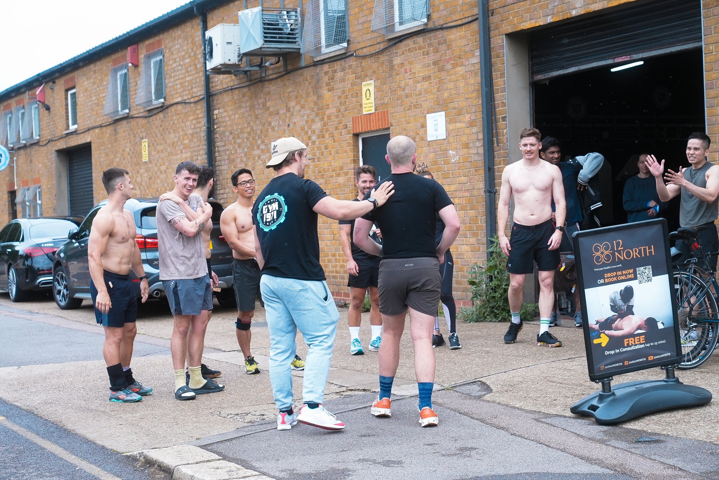 Group of men, some shirtless, gather outside a brick building, laughing and chatting.