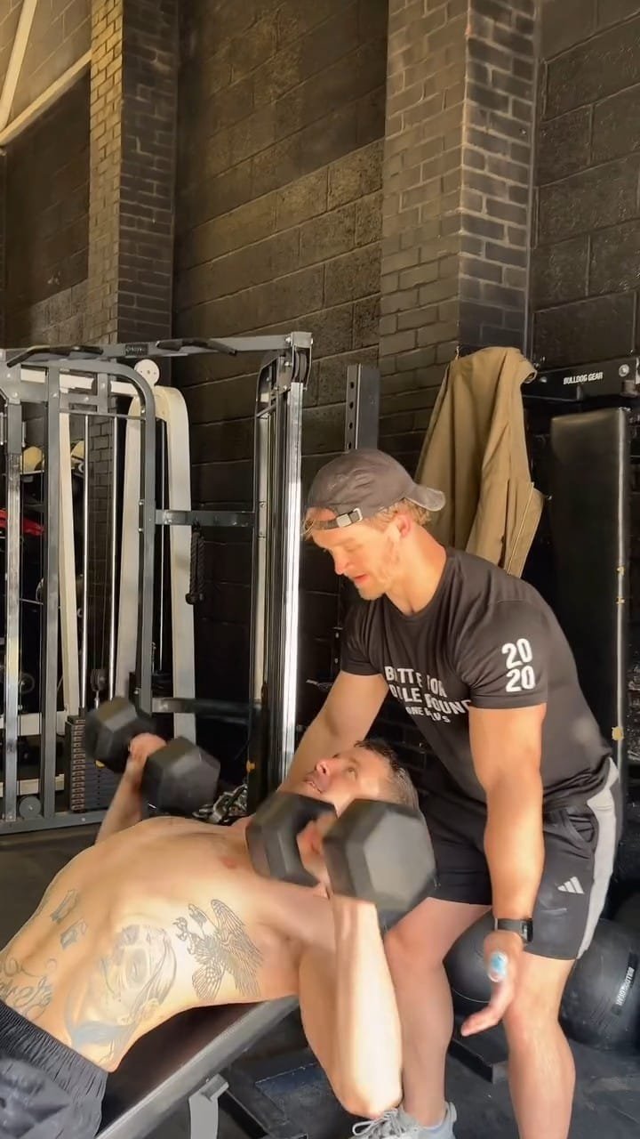 A man lying on a gym bench lifting dumbbells with the assistance of a trainer in a gym with brick walls.