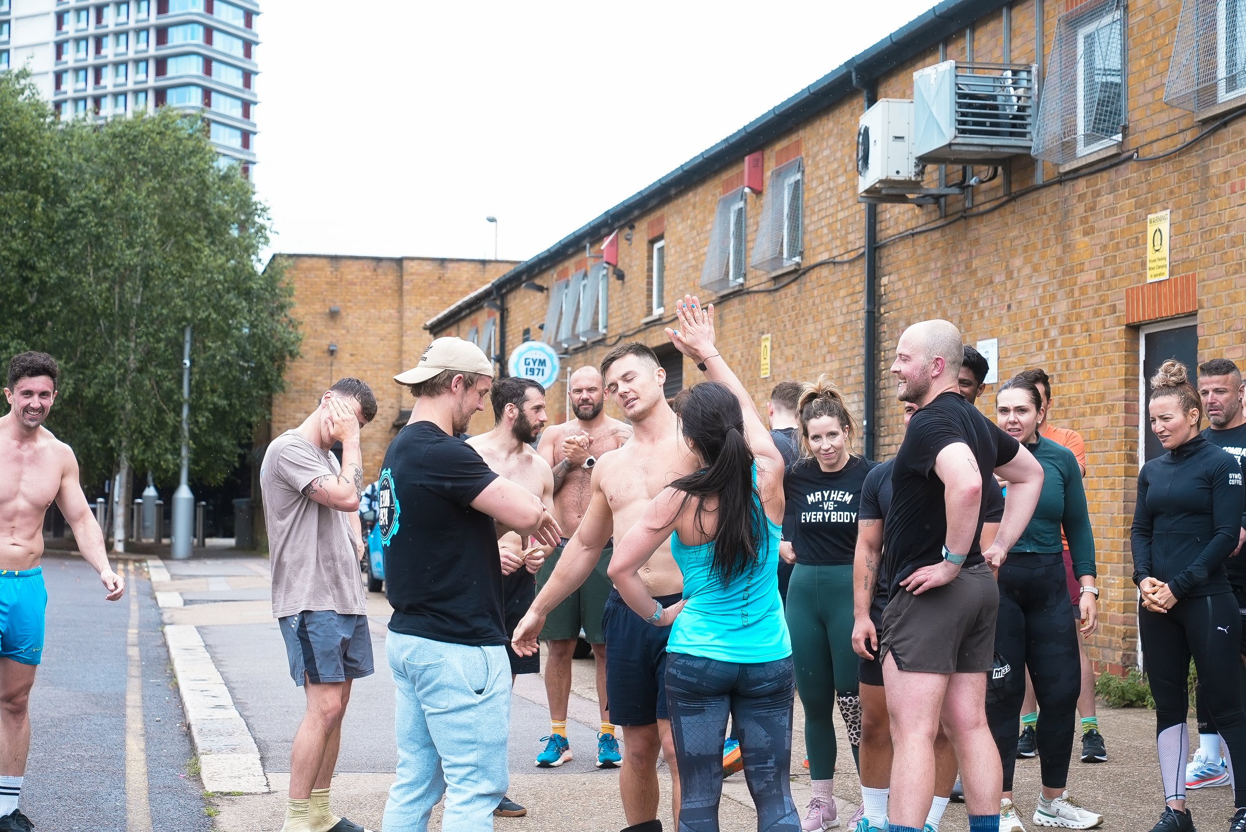 A group of people gathered outdoors beside a brick building, with some shirtless and wearing workout clothes, engaging in a fitness class or workout session, after which they are socializing and smiling.
