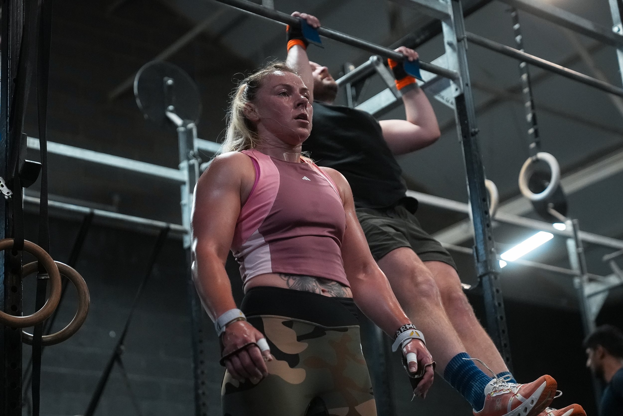 A woman in athletic clothing and gloves, sweating and focused, stands in a gym next to a man hanging from a pull-up bar, both preparing for or finishing a workout.