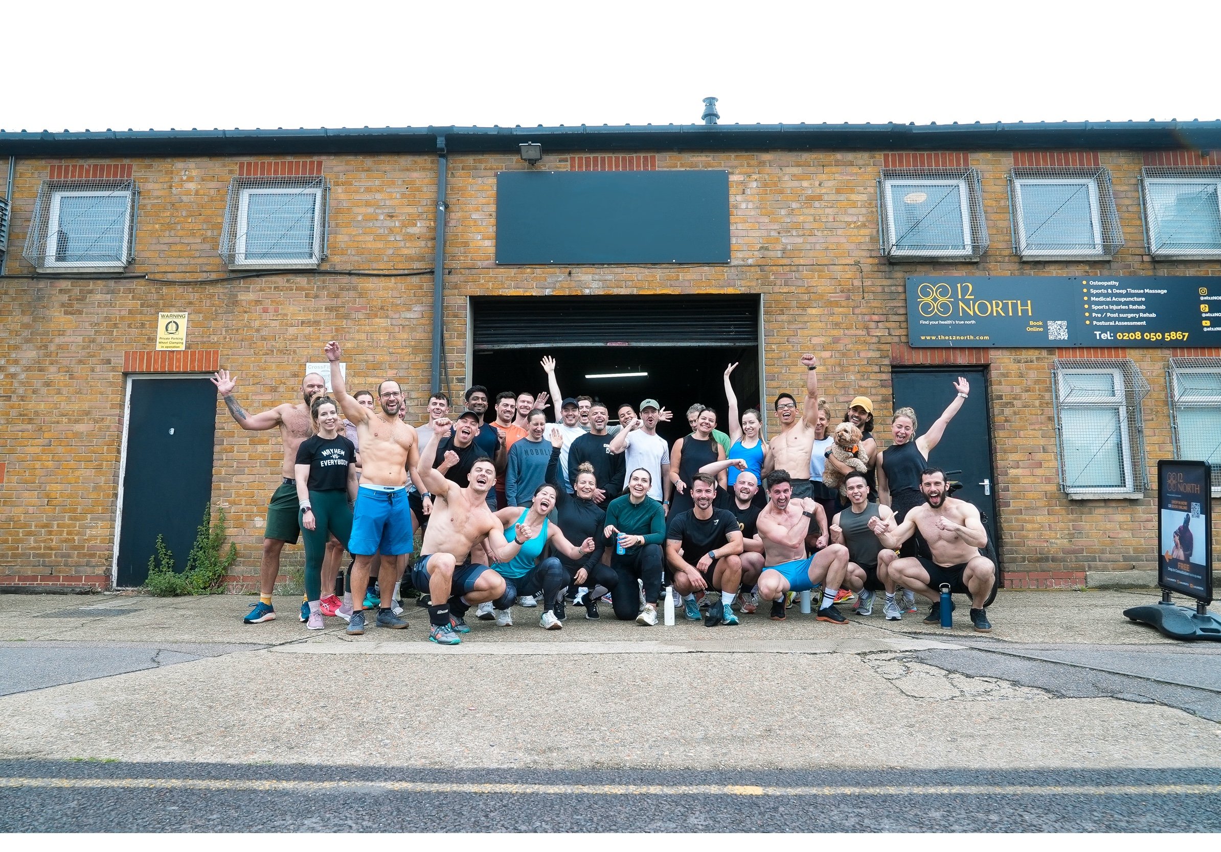 Group of people celebrating outside a gym in front of a brick building, some shirtless and wearing athletic clothes, with a sign that reads 'GYM 1971' above the entrance.