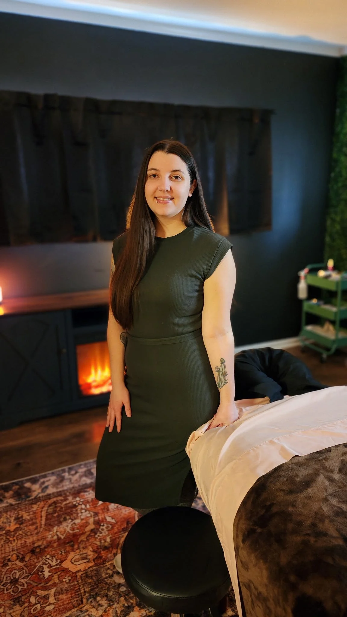 A woman in a dark green dress standing beside a bed in a dimly lit room with a fireplace, black curtains, and green wall decor.