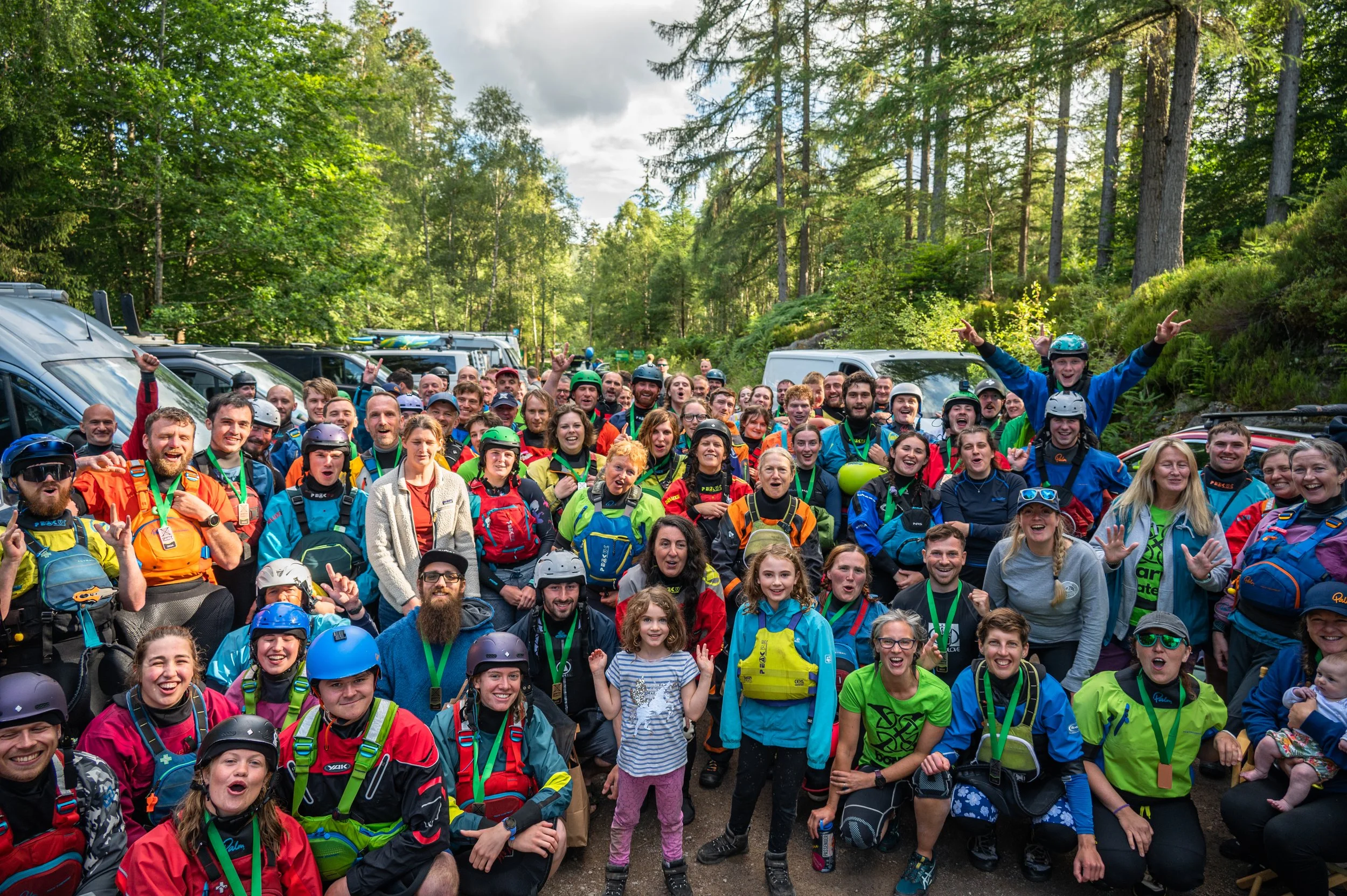 Group photo of smiling people in outdoor gear, including helmets and backpacks, gathered in a forested area for a group event or adventure.