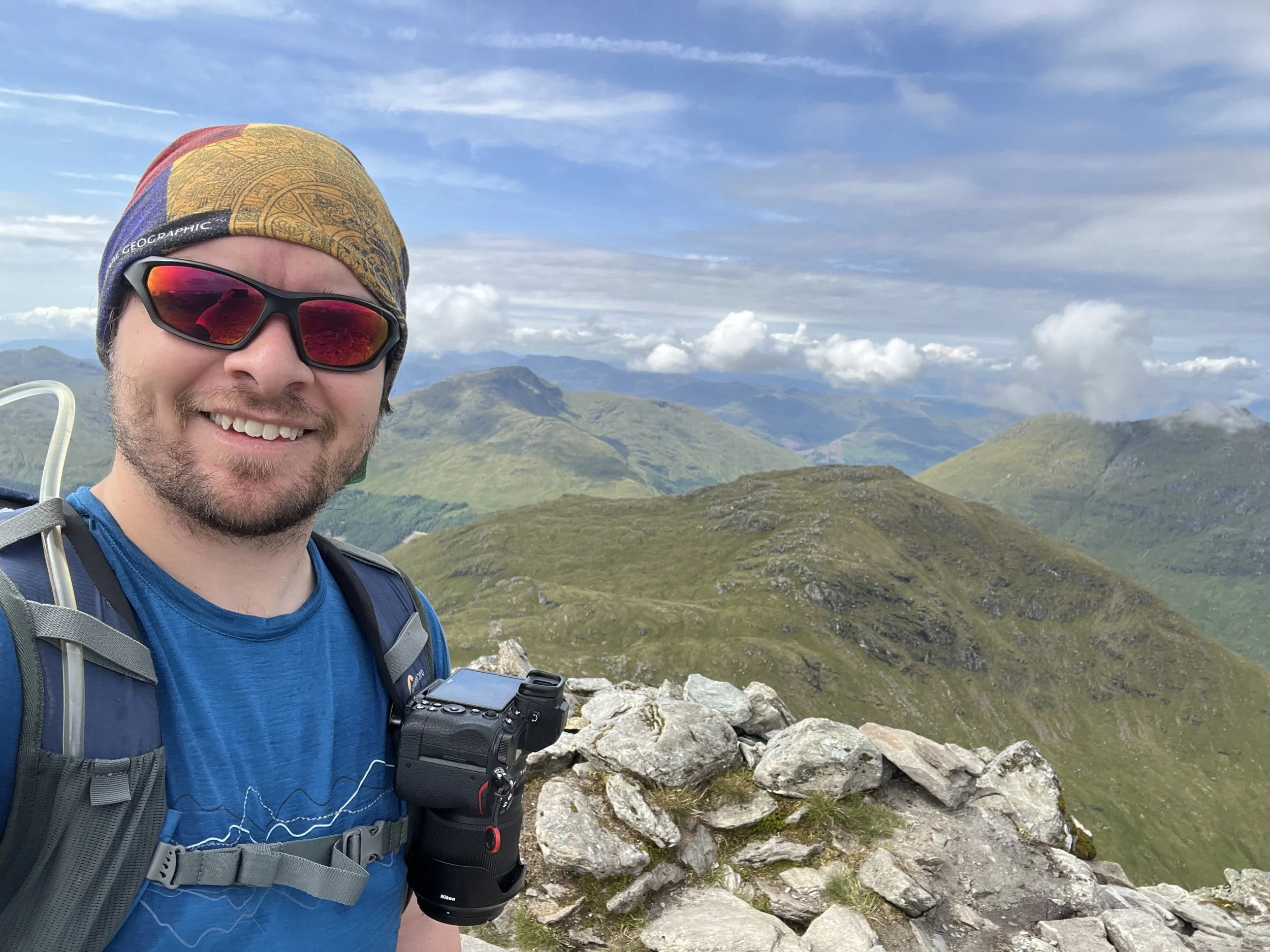 Man in blue shirt with a backpack and camera, smiling, on a mountain trail with green hills and cloudy sky in the background.