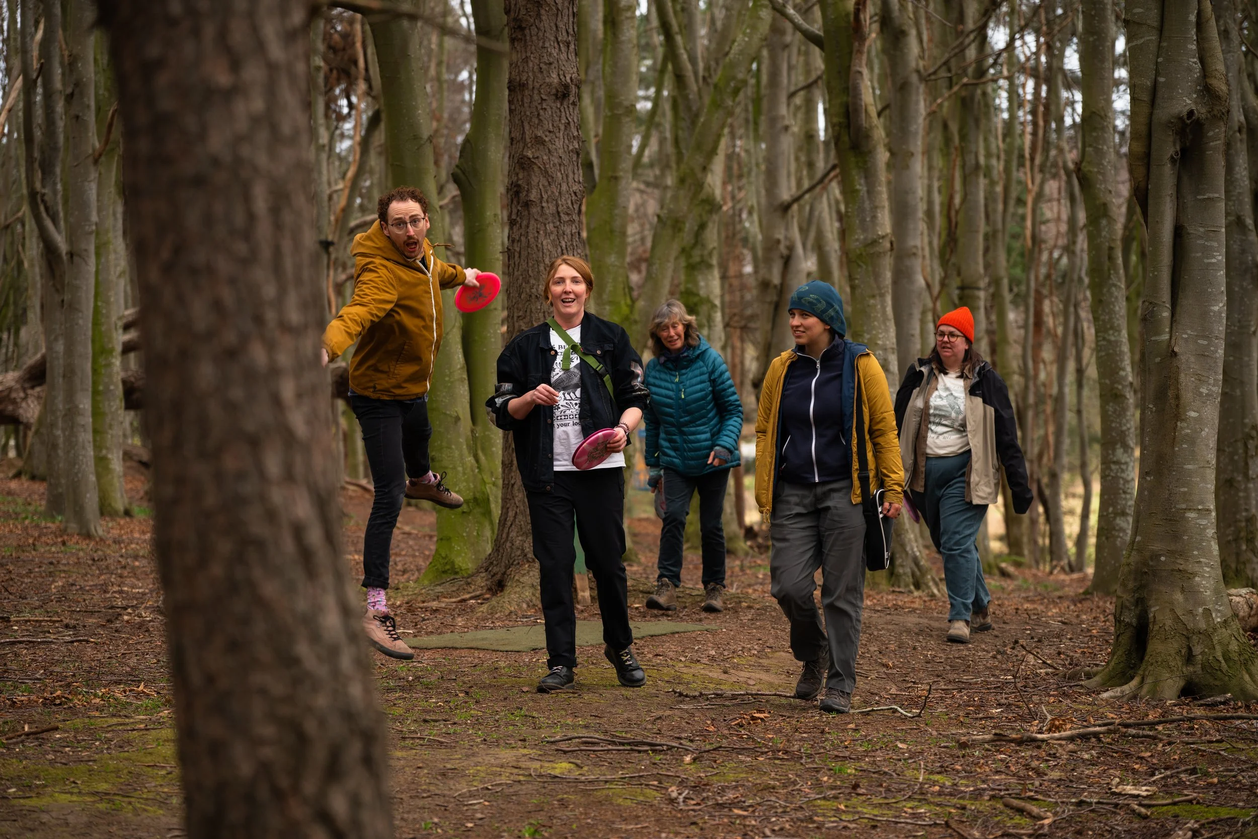 A group of five people enjoying a walk in a forest, with one person jumping and holding a pink frisbee, all smiling and dressed in casual outdoor clothing.