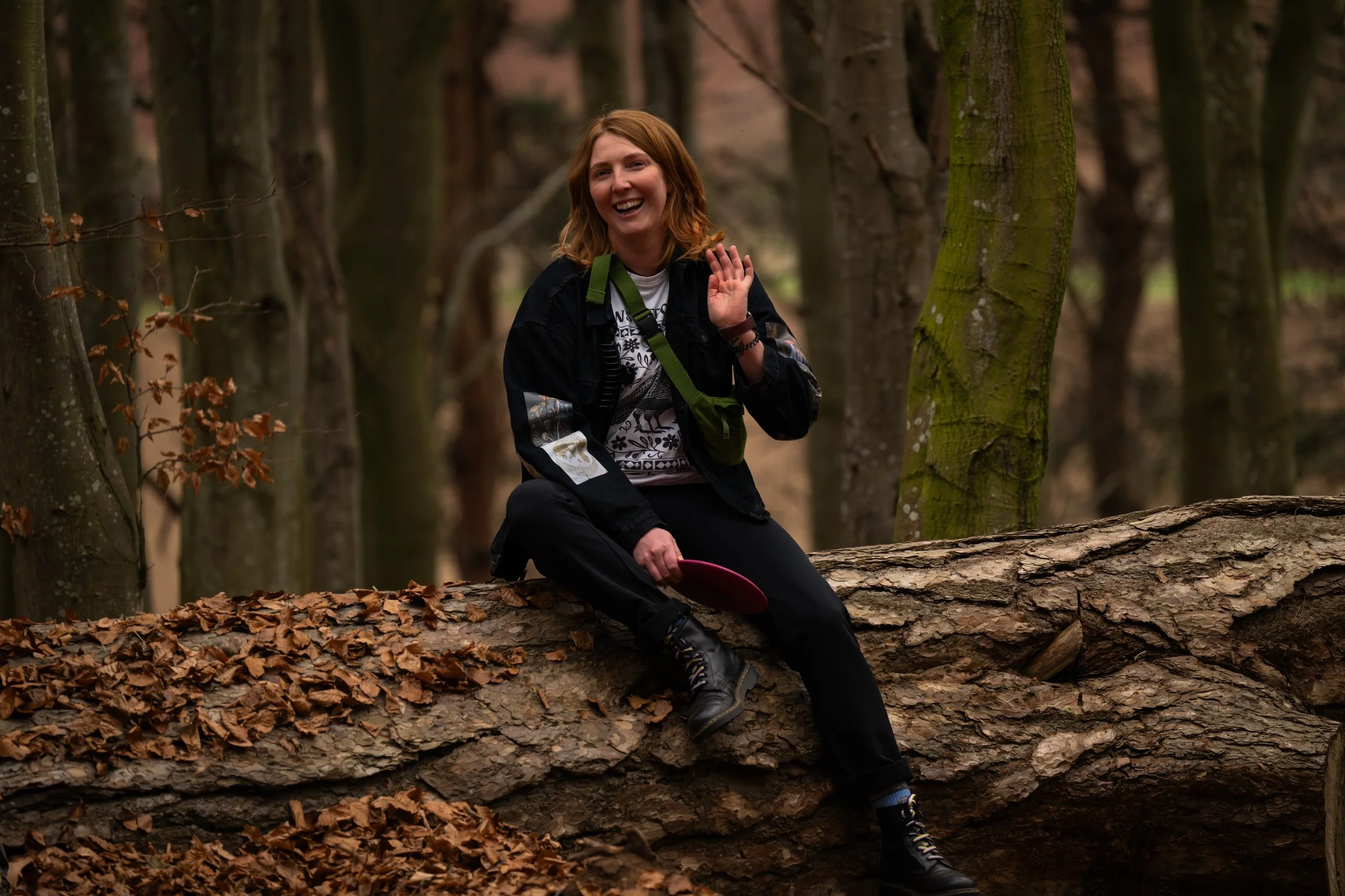 A woman with red hair sitting on a fallen tree in a forest, smiling and waving, with a small green bag across her chest and holding a pink frisbee.