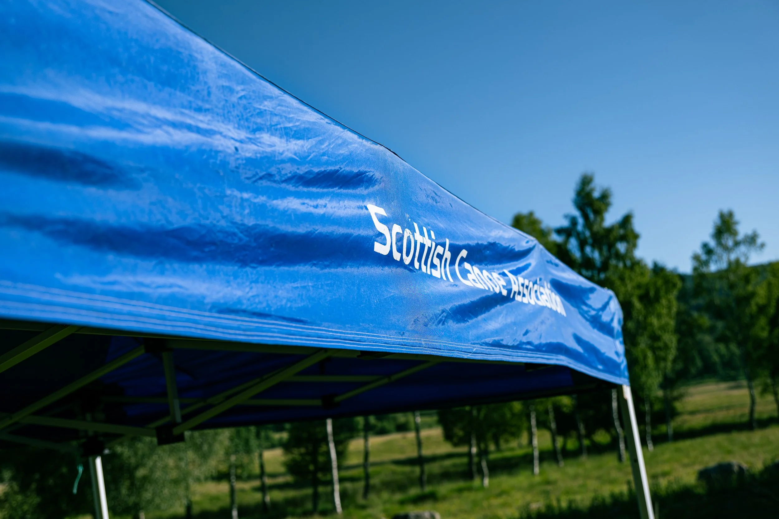 Close-up of a blue tent with 'Scottish Canoe Association' written on it, set outdoors with trees and a clear blue sky in the background.