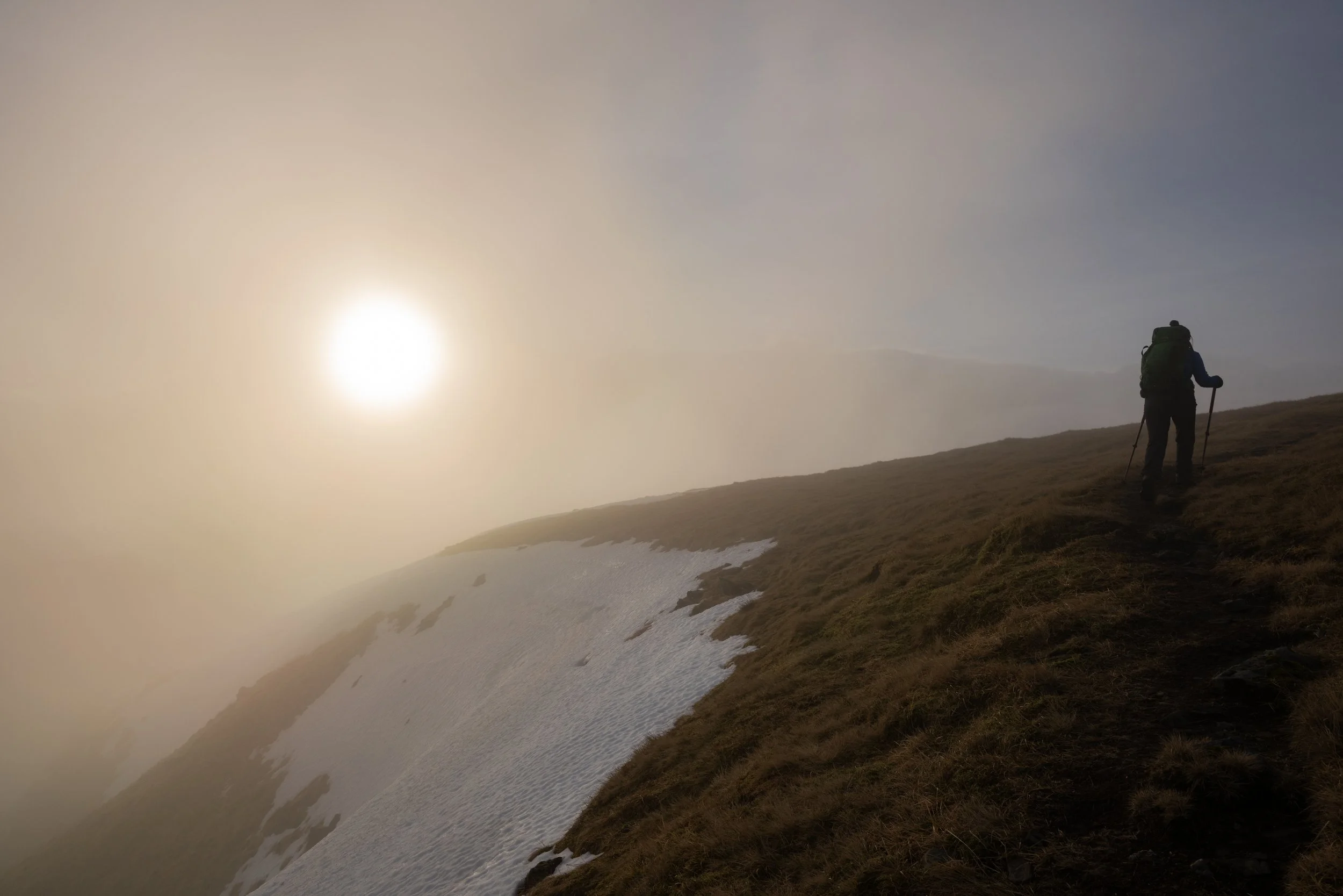 Hiker in outdoor gear climbing a hill on a narrow trail, with snow patches on the ground and a misty sky in the background, illuminated by a hazy sun.