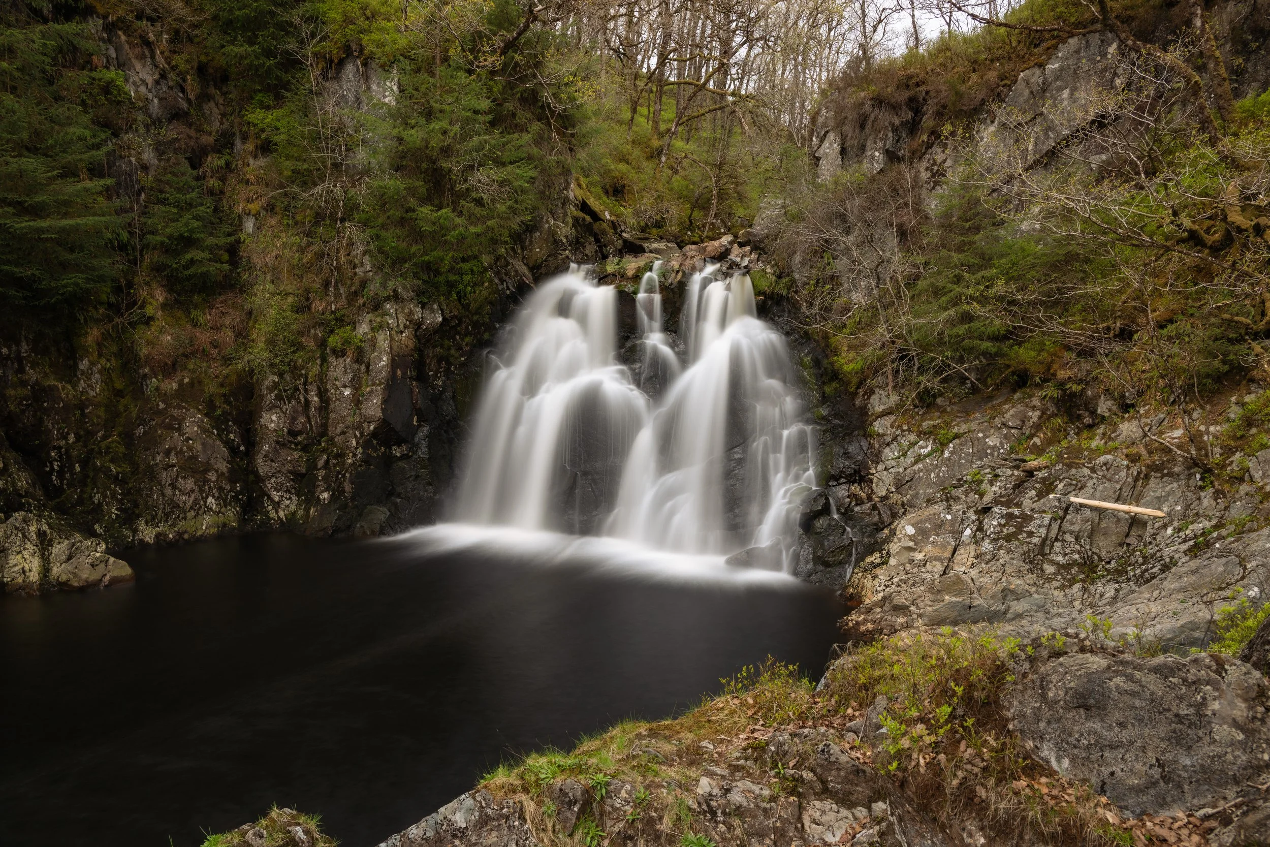 A waterfall cascading into a dark pool surrounded by rocky terrain and lush green trees in a forest.