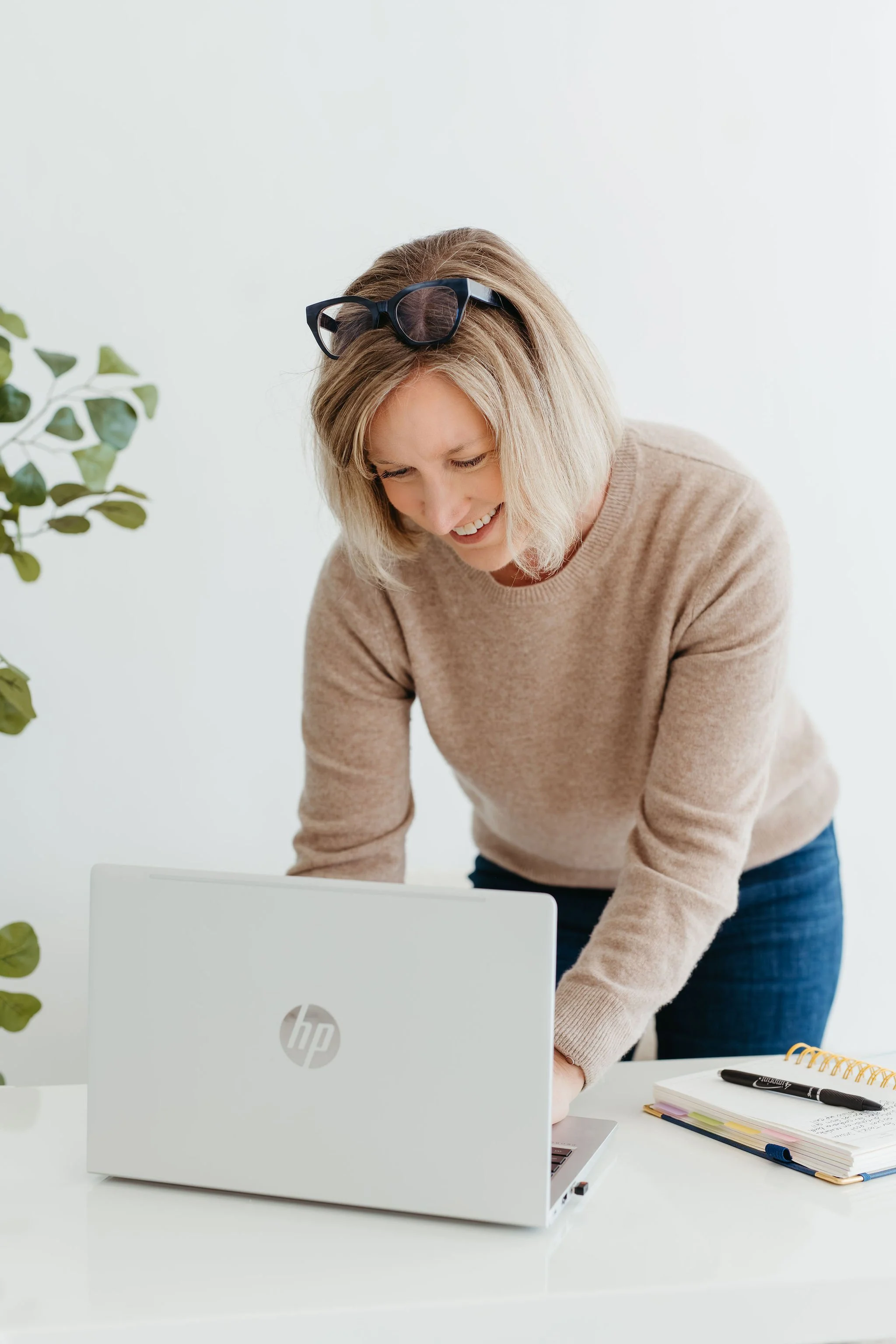 A woman with blond hair wearing sunglasses on her head, smiling and leaning over a white desk with a laptop, notebooks, a pen, and a plant nearby.