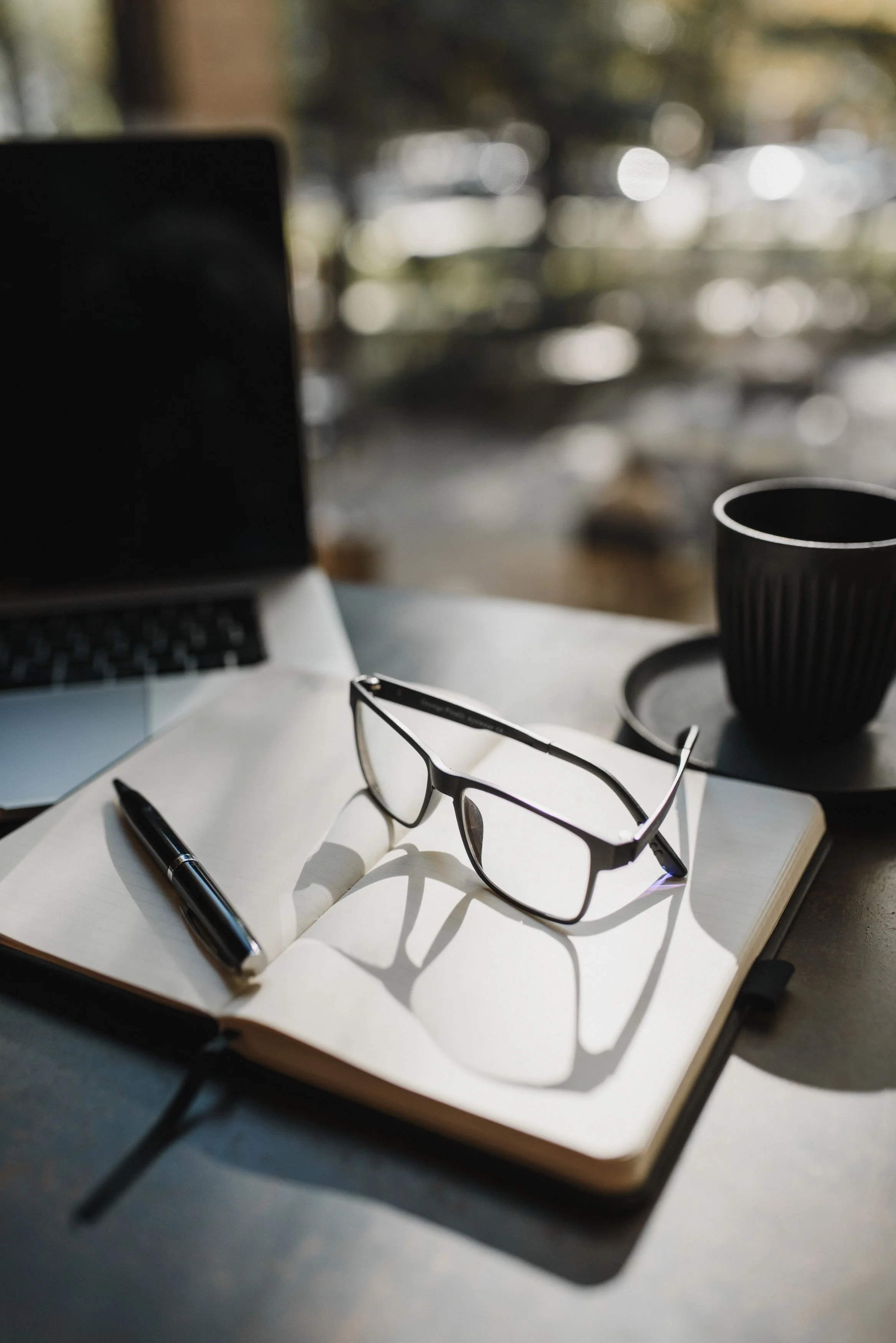 A workspace with a black pen, a pair of eyeglasses, a spiral notebook, and a black ceramic mug on a table. In the background, a laptop is partially visible, and the scene is illuminated by natural light.