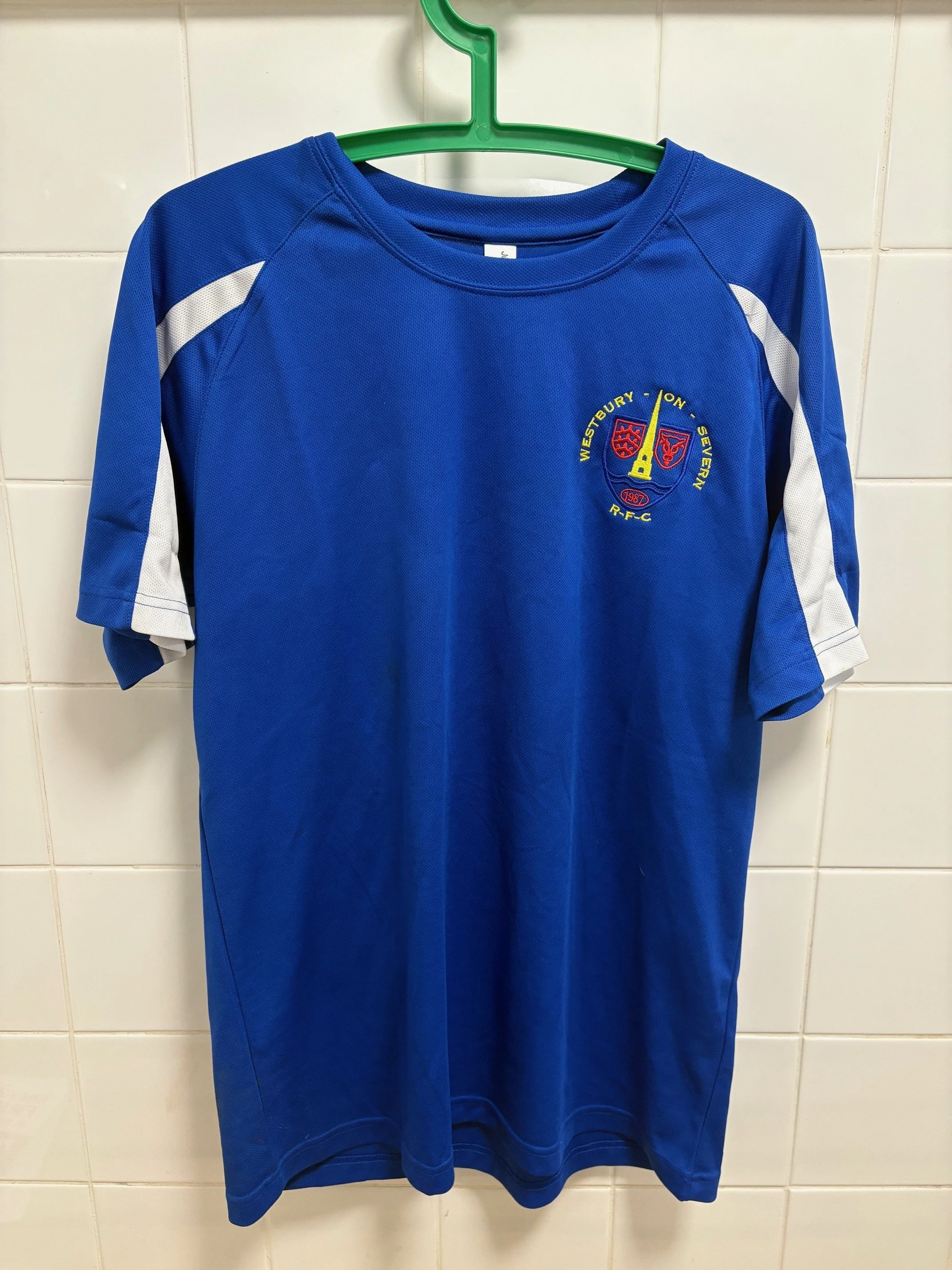 Blue sports jersey with white accents on sleeves and a crest embroidered on the chest, hanging on a green hanger against a tiled wall.