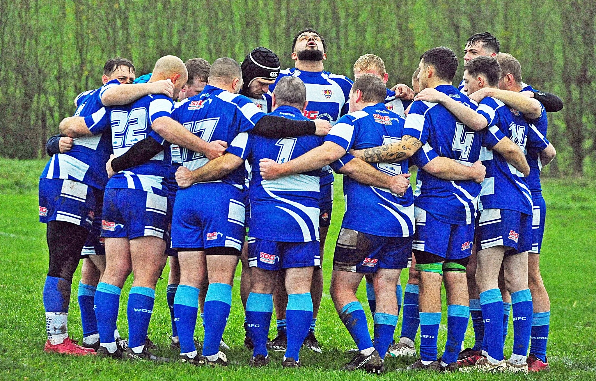 A group of rugby players wearing blue and white uniforms huddled together on a grassy field, with one player in the center looking upward.