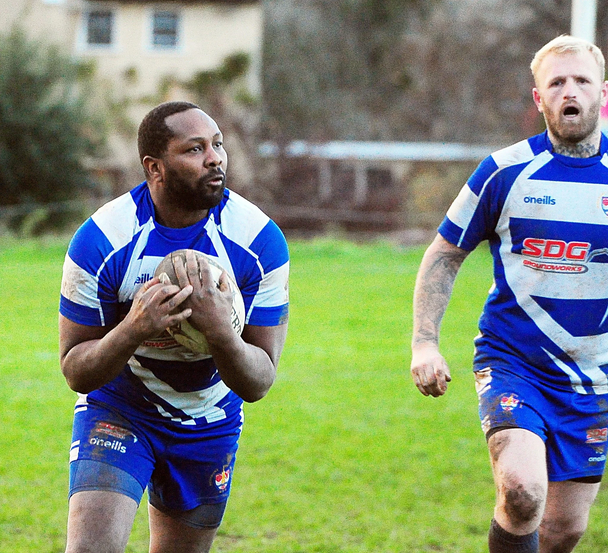 Two rugby players in blue and white uniforms during a match, one holding a rugby ball and the other running beside him.