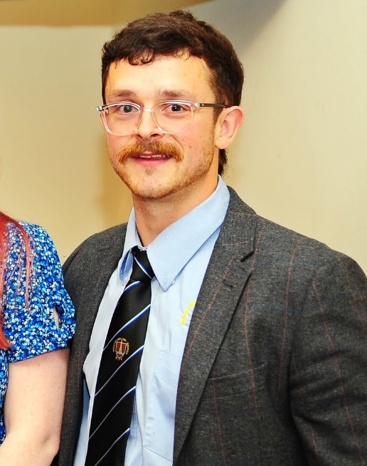 A man with a beard and messy hair wearing a vest, dress shirt, and striped tie, smiling at the camera.