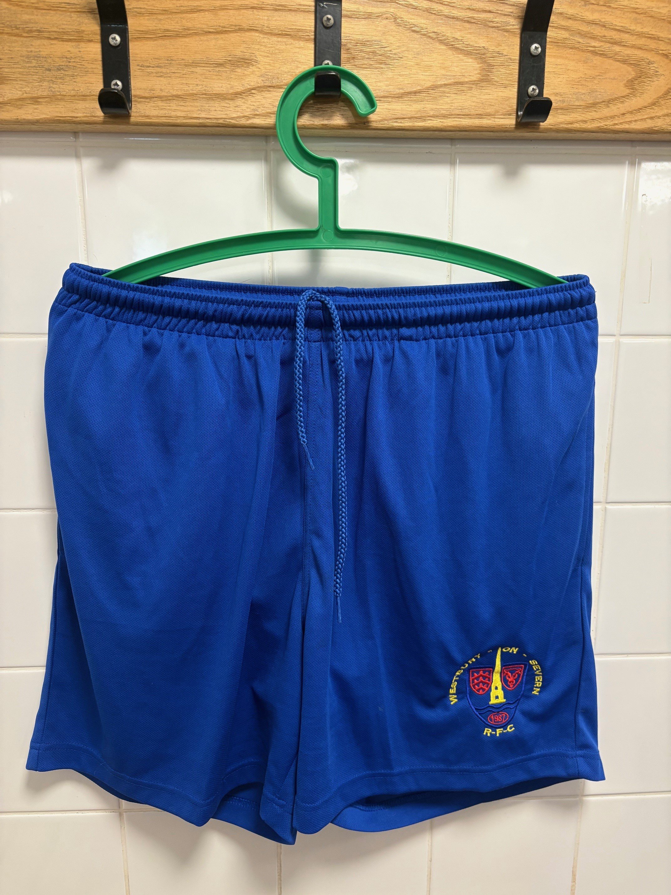 Blue sports shorts with a logo hanging on a green hanger against a tiled wall.
