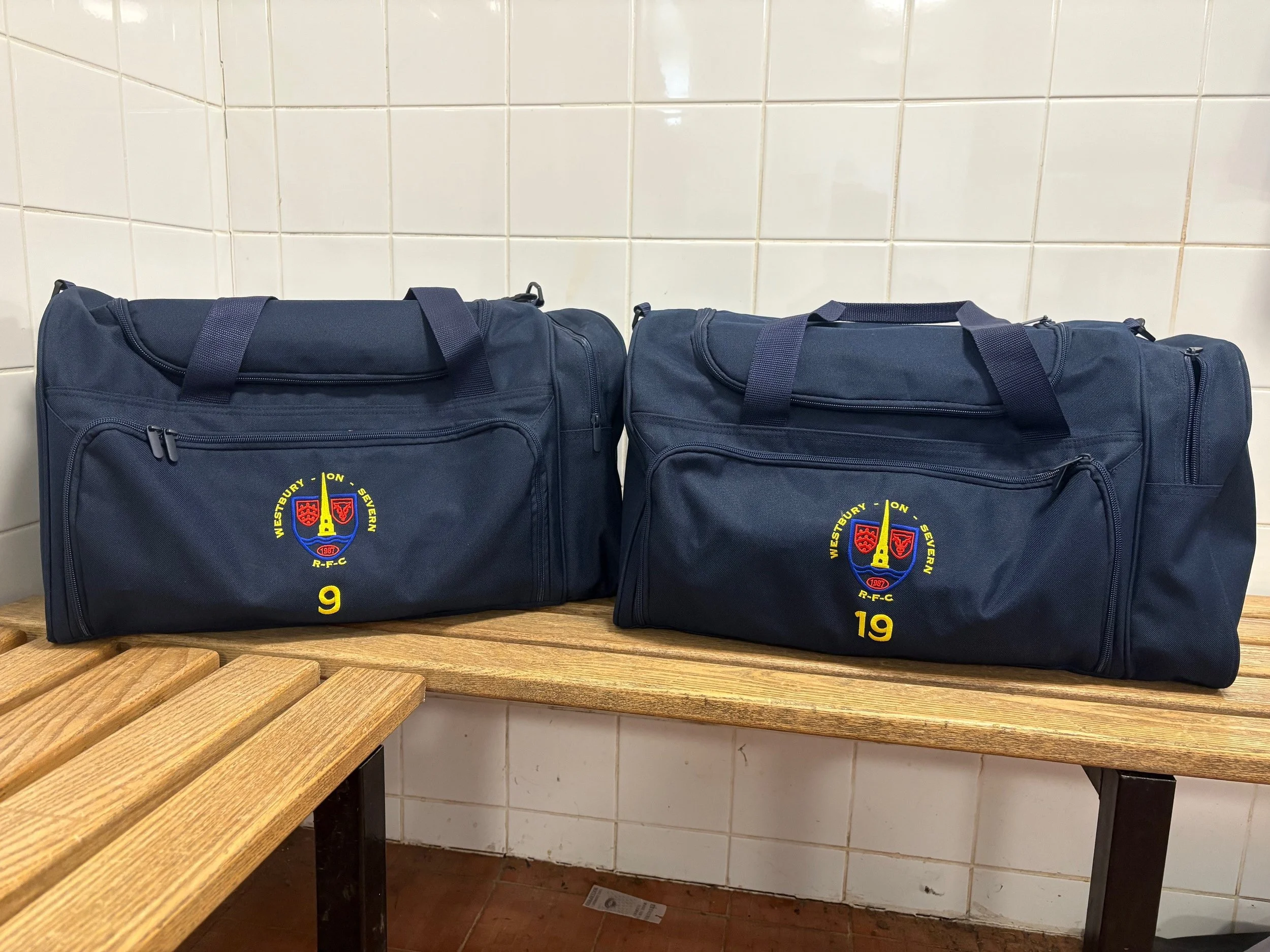 Two navy blue sports bags with team emblem placed on a wooden bench against a white tiled wall.
