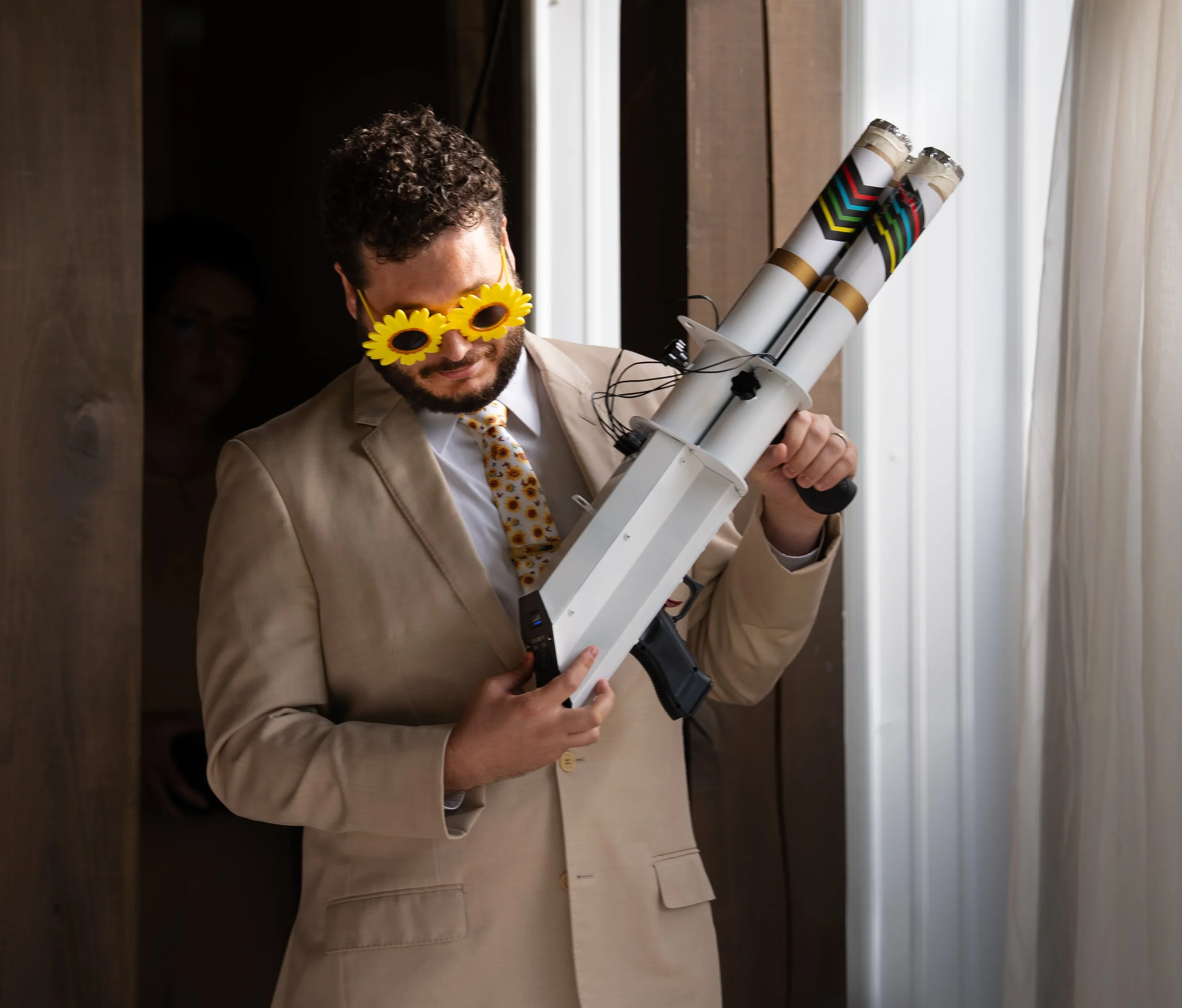 Man with curly hair and beard wearing sunflower sunglasses, beige suit, and floral tie, holding a rocket launcher with rolled-up posters, smiling by a window.