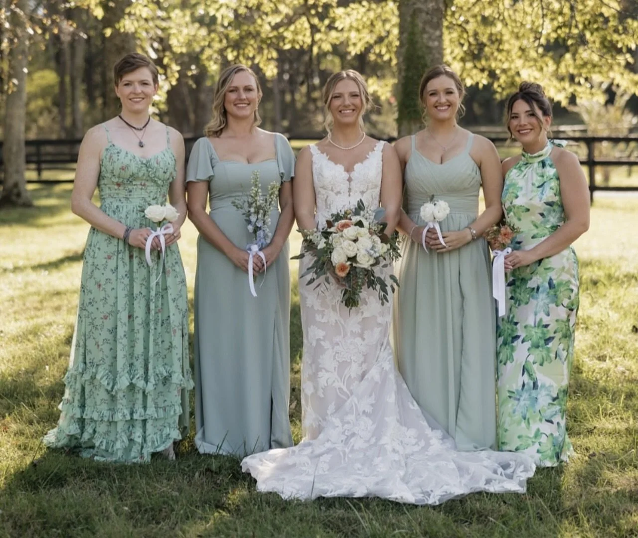 Group of five women standing outdoors on a sunny day, dressed in formal dresses, with trees in the background. One woman in the center is wearing a white wedding gown and holding a bouquet, surrounded by four women in pastel-colored dresses, each hol