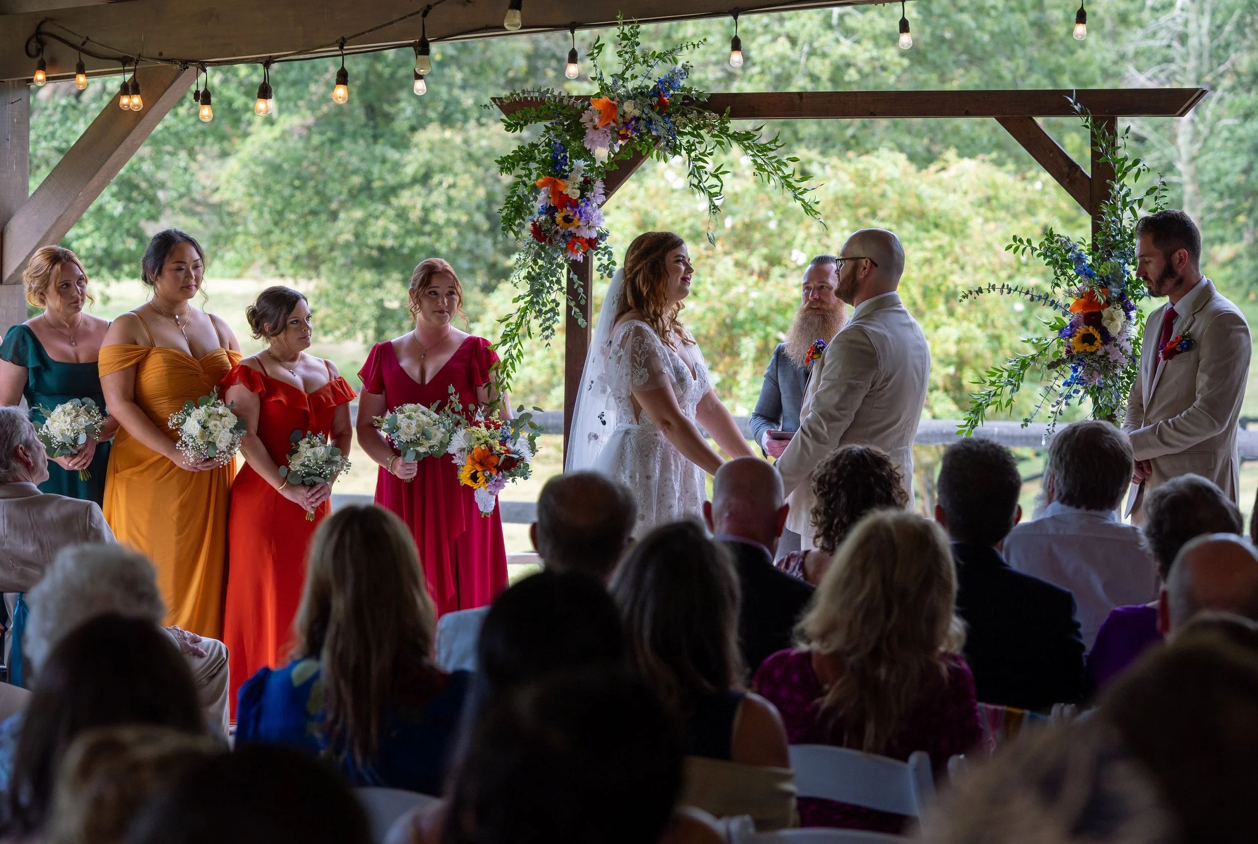 A wedding ceremony with a bride and groom holding hands under a decorated arch, surrounded by bridesmaids and groomsmen, in an outdoor setting with green trees in the background, during daytime.