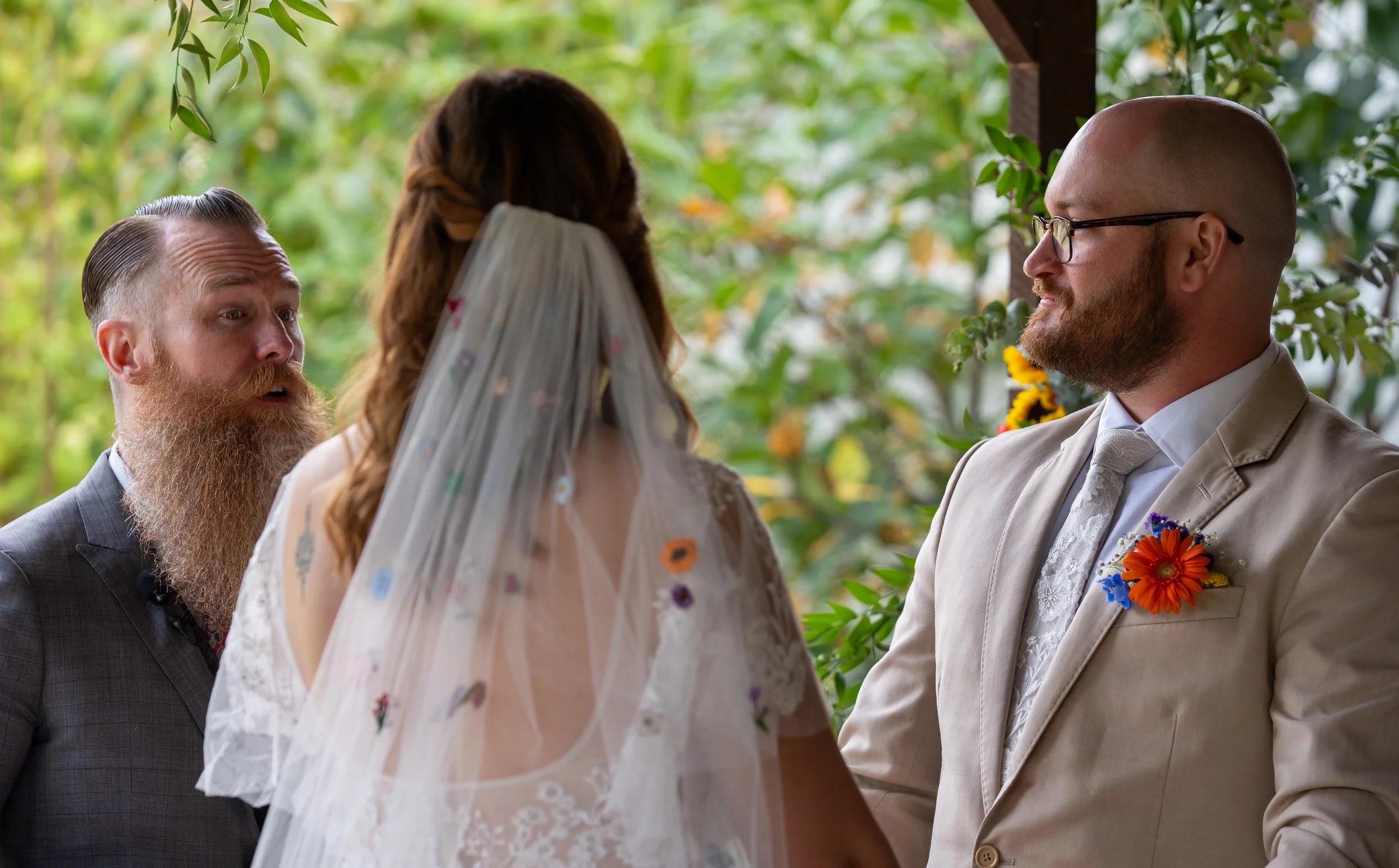 A couple getting married outdoors, with officiant and officiant's assistant, all in formal attire and floral accessories, surrounded by green foliage.
