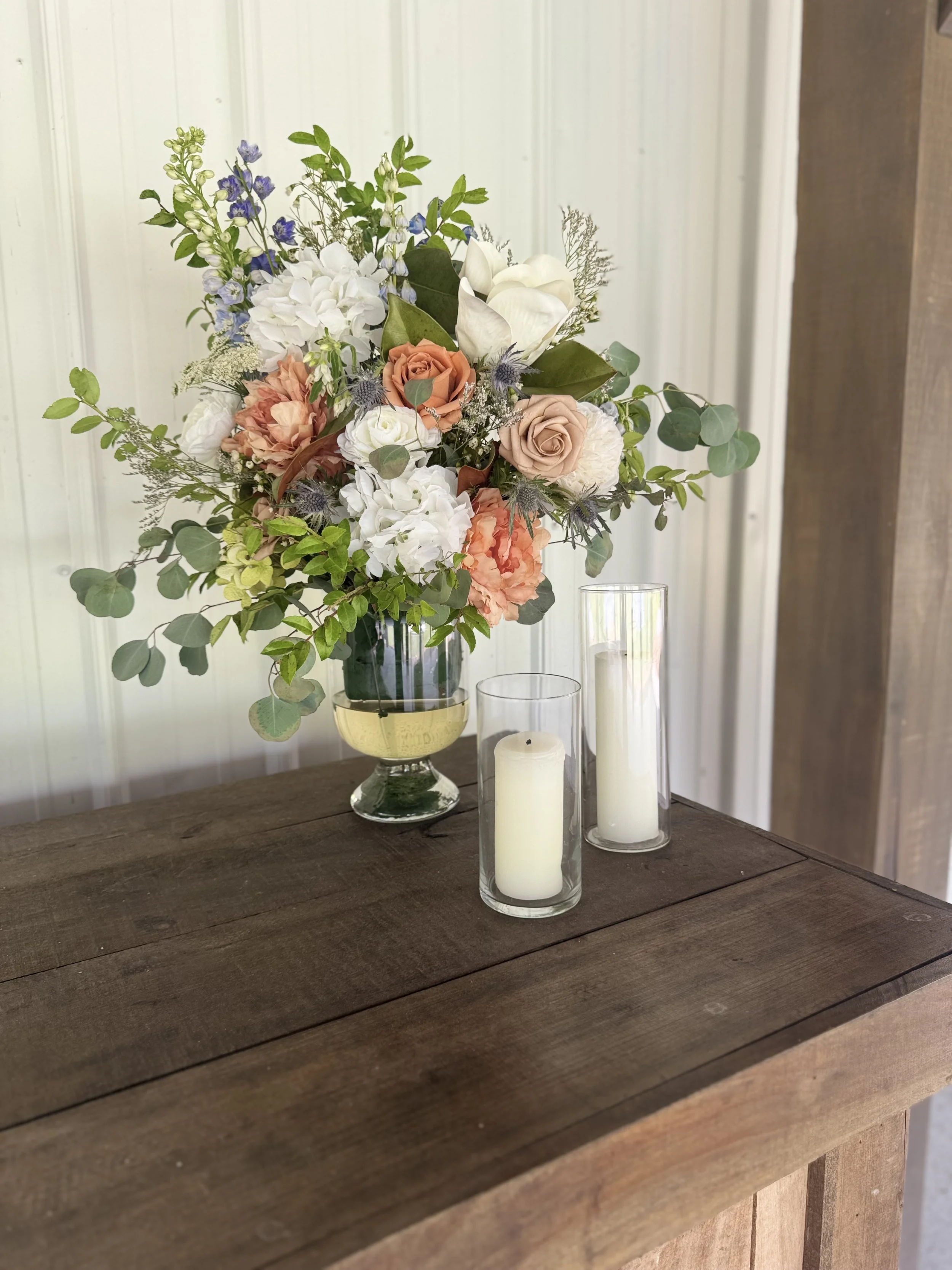 A floral arrangement with white, peach, and blue flowers and green leaves in a dark vase, flanked by two white candles in glass holders, on a wooden table against a white paneled wall.