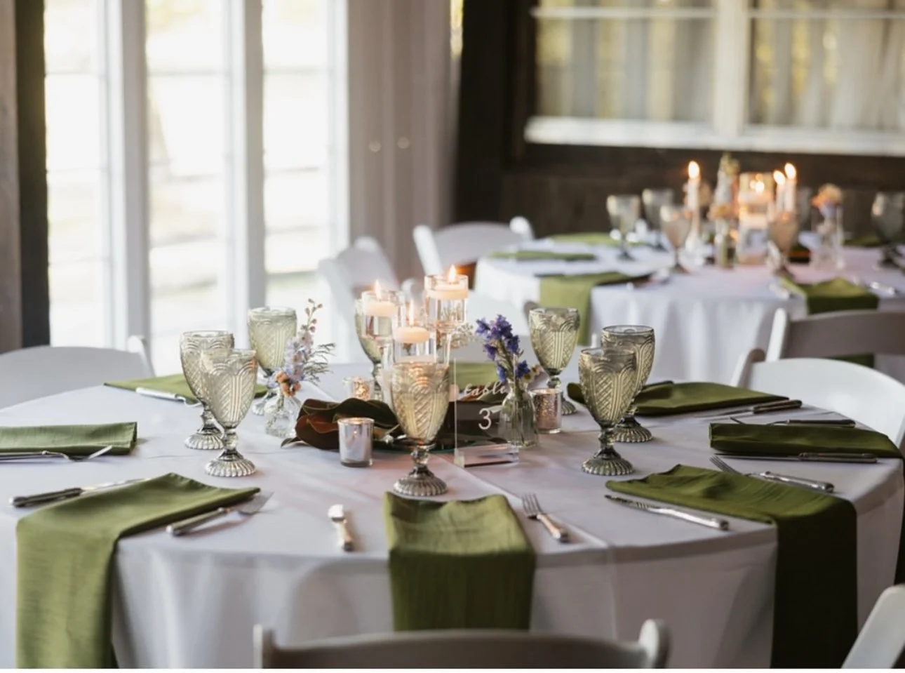 Table set for a formal event with white tablecloths, green napkins, vintage-style glassware, floral centerpieces, and lit candles in a warmly lit room with large windows.