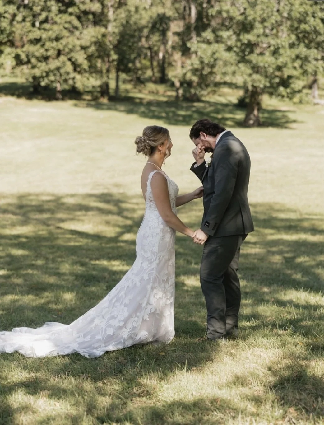 A bride and groom are holding hands outdoors on a grassy field, sharing an emotional moment with the groom wiping away tears. The bride is wearing a white lace wedding dress with a train, and the groom is in a gray suit. Trees with green foliage are