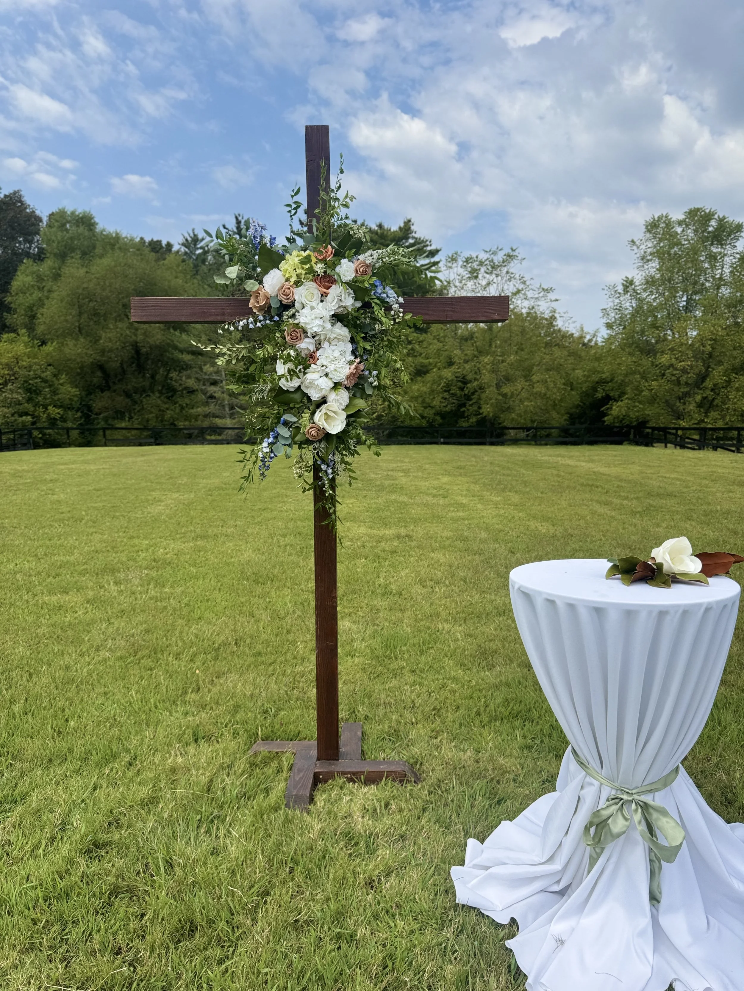 An outdoor wedding setup with a wooden cross decorated with flowers and greenery, and a small round table covered with white fabric and a ribbon, with flowers on top. The setting is on a grassy field surrounded by trees under a partly cloudy sky.