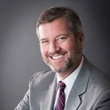 A smiling man with a beard and gray hair, wearing a light gray suit jacket, white dress shirt, and striped tie, against a gray background.