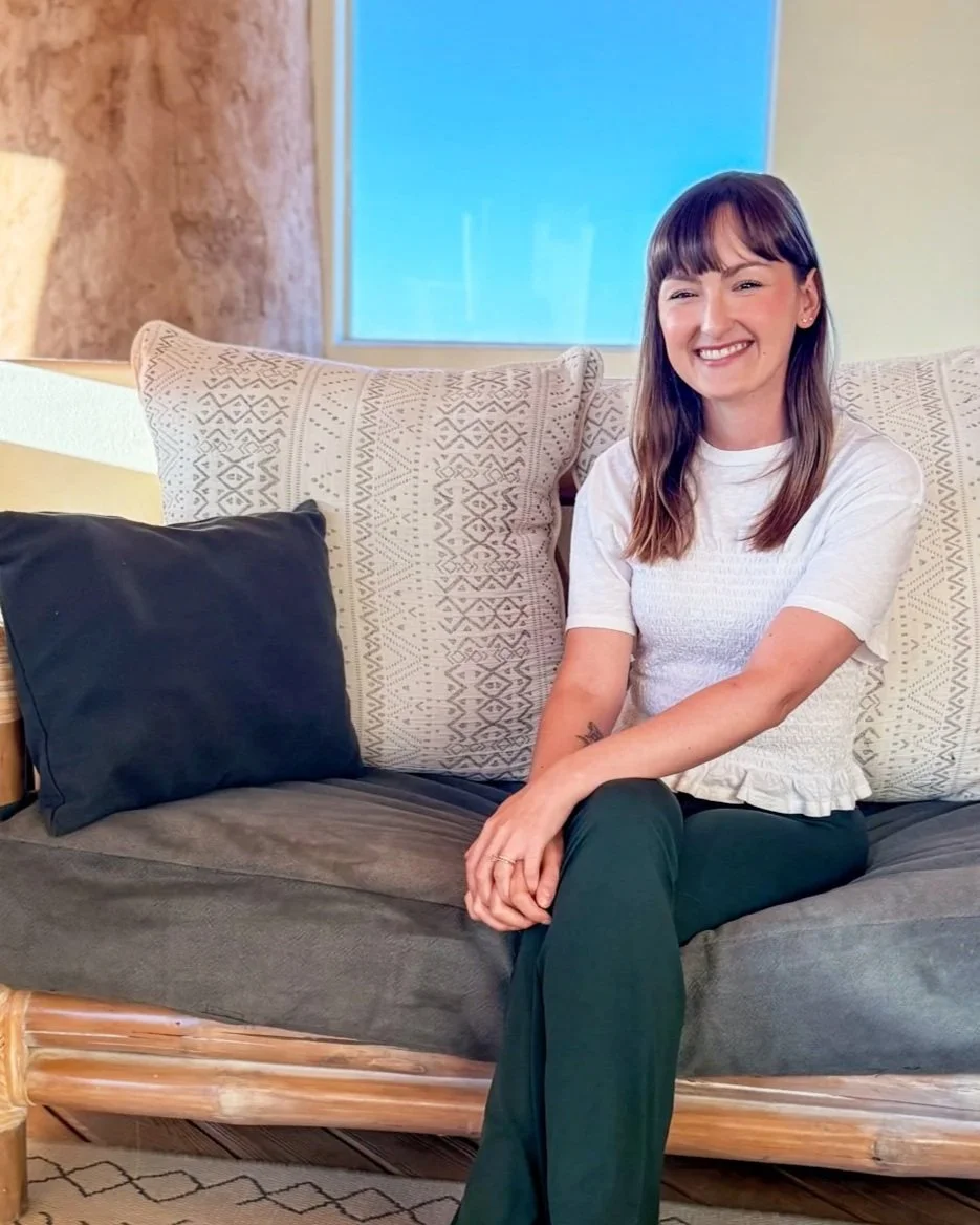 A woman sitting on a sofa with a light-colored patterned pillow, smiling, with a dark pillow beside her and a window in the background.