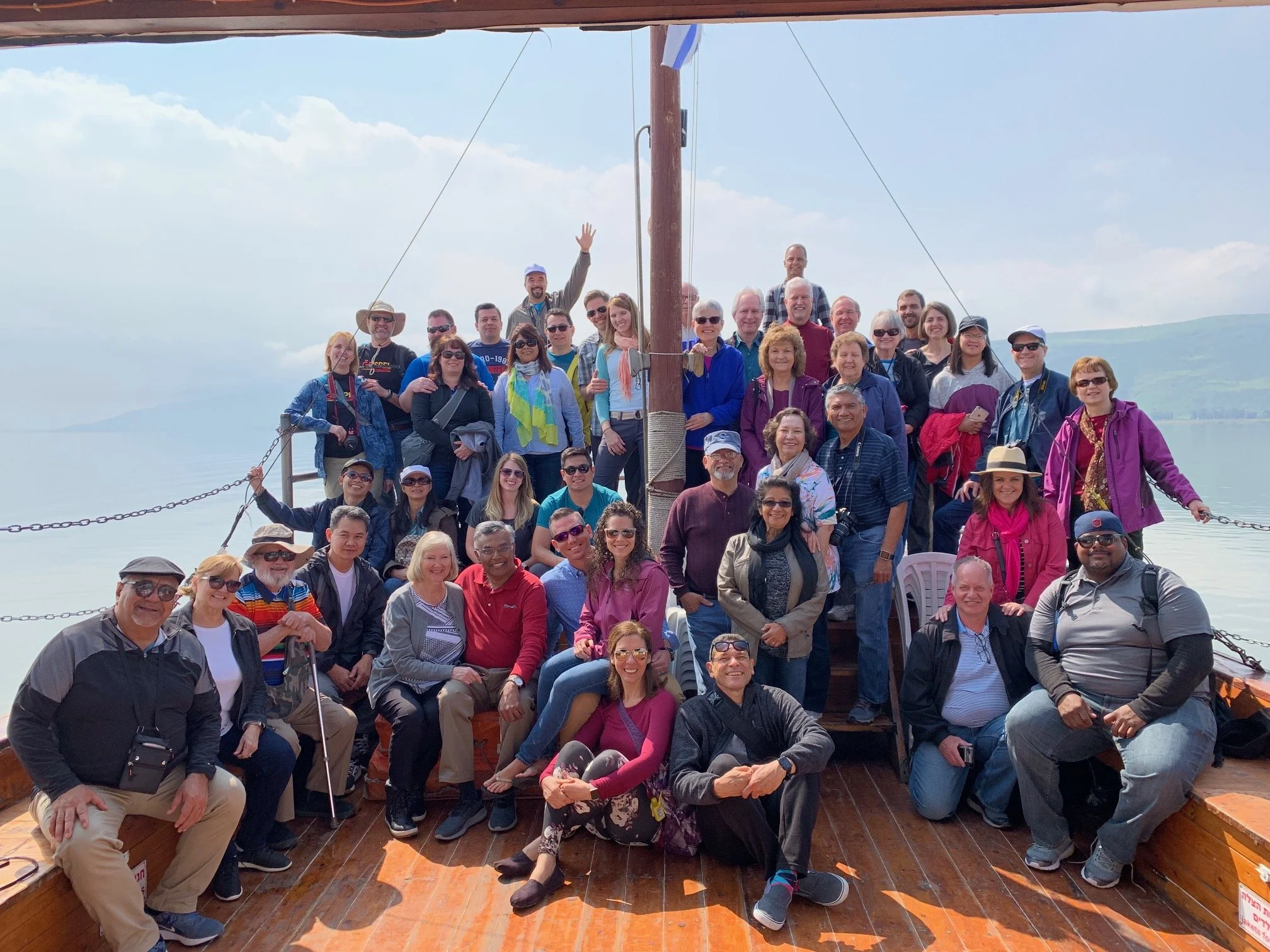Group of diverse people on a boat cruise, smiling and posing for a photo with a water and mountainous landscape in the background.