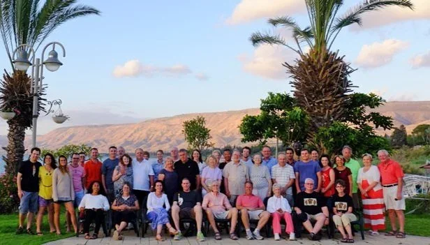 Group of people outdoors in front of palm trees, with mountains and a blue sky in the background.