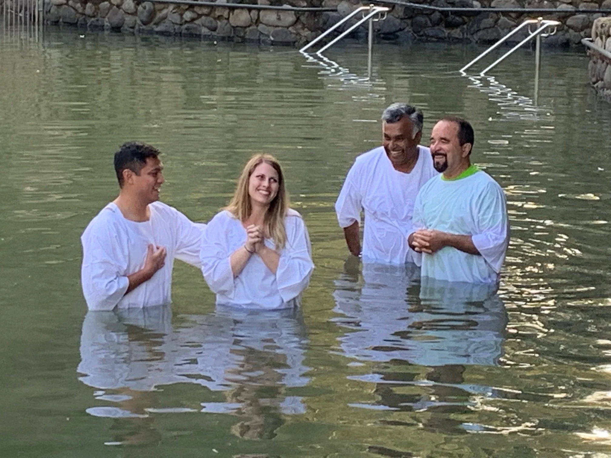 Four people standing in a body of water, possibly during a religious ceremony or baptism, wearing white robes and smiling.