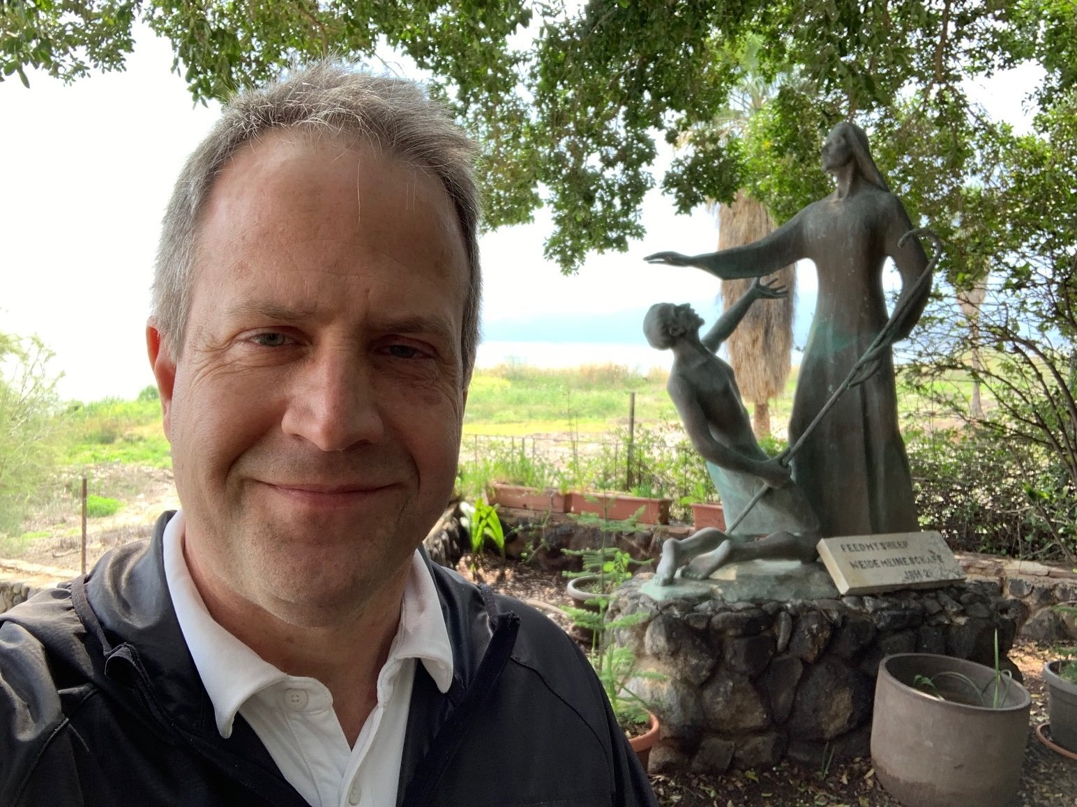 A man taking a selfie outdoors with a sculpture of two women and a child in the background. The sculpture is on a rock base, and there are plants and trees around.