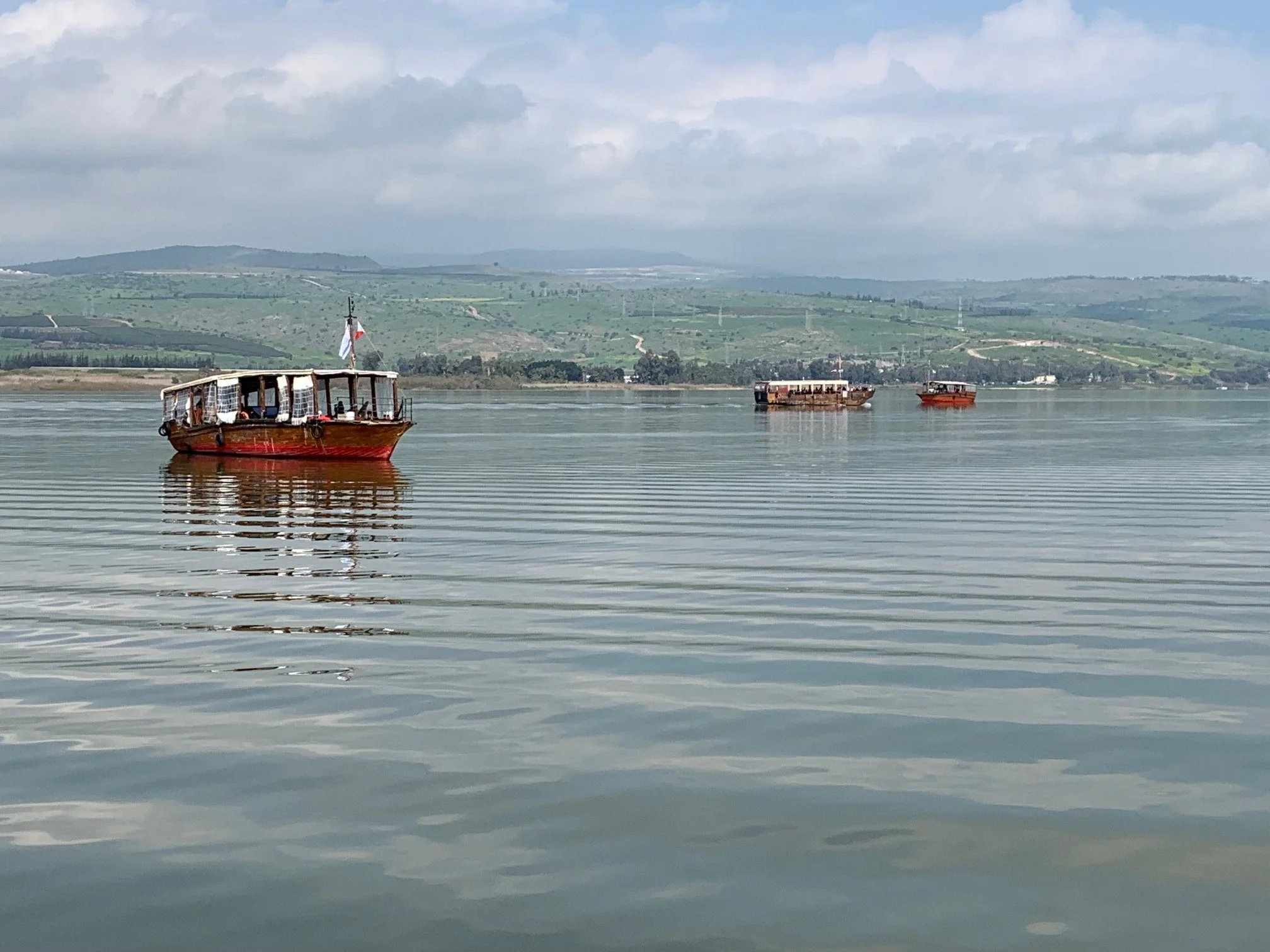 Three boats on a calm body of water with green hills and a cloudy sky in the background.