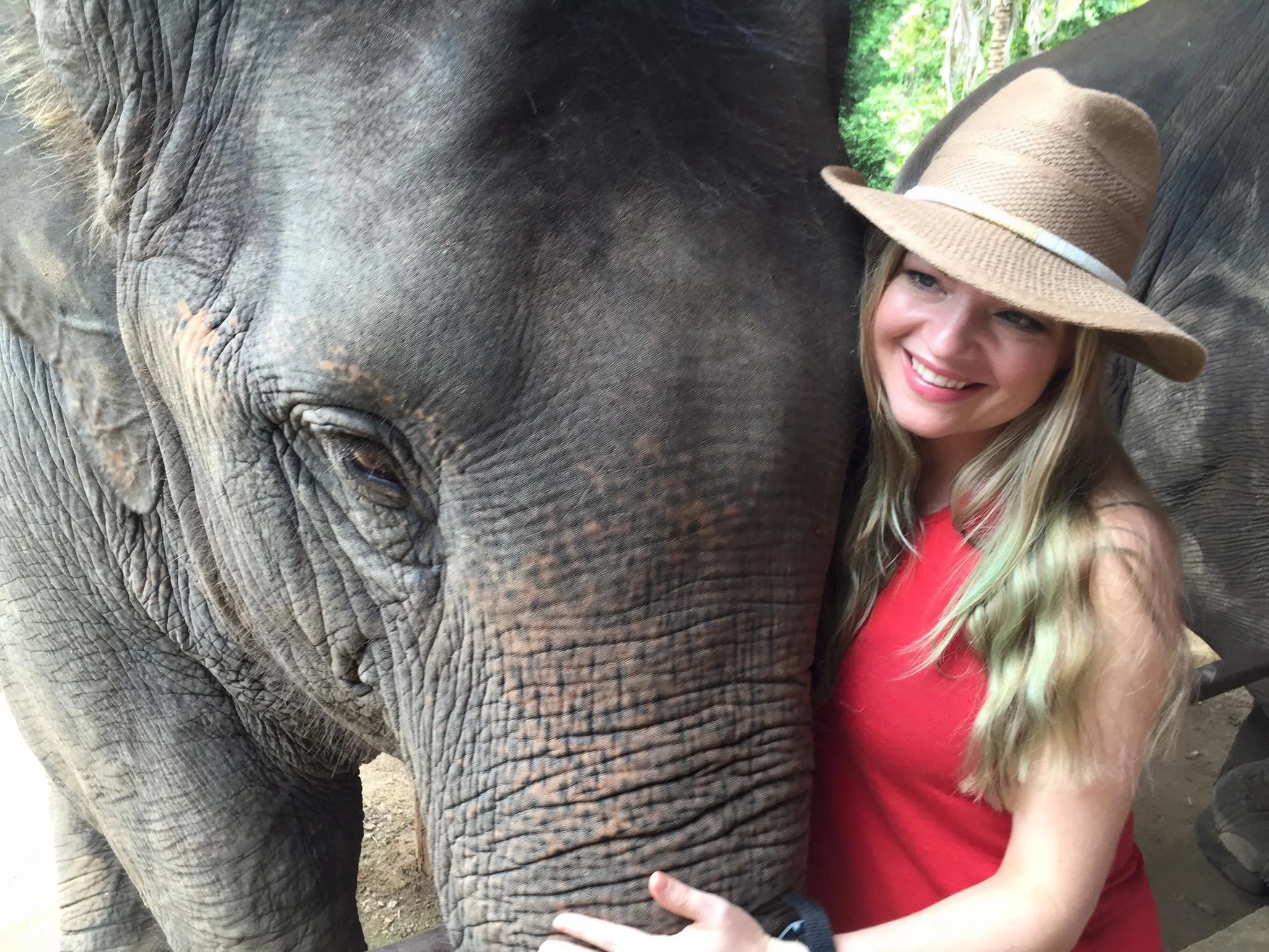 A woman with long blonde hair, wearing a beige sunhat and a red sleeveless top, hugging an elephant and smiling, with green trees in the background.