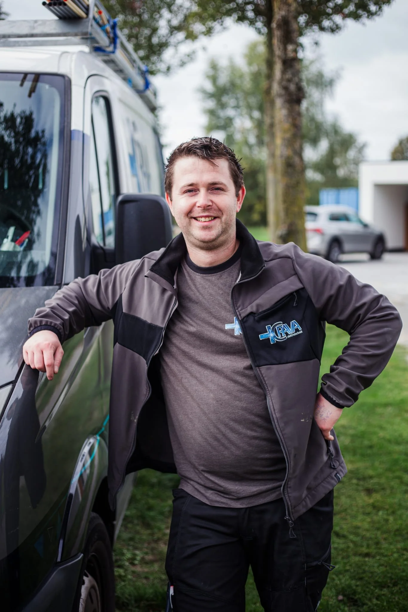 Een man met kort, donker haar die lacht, aangetrokken in een grijze en zwarte jas met logo, staande naast een auto met een ladder op het dak, in een tuin met bomen en een huis op de achtergrond.