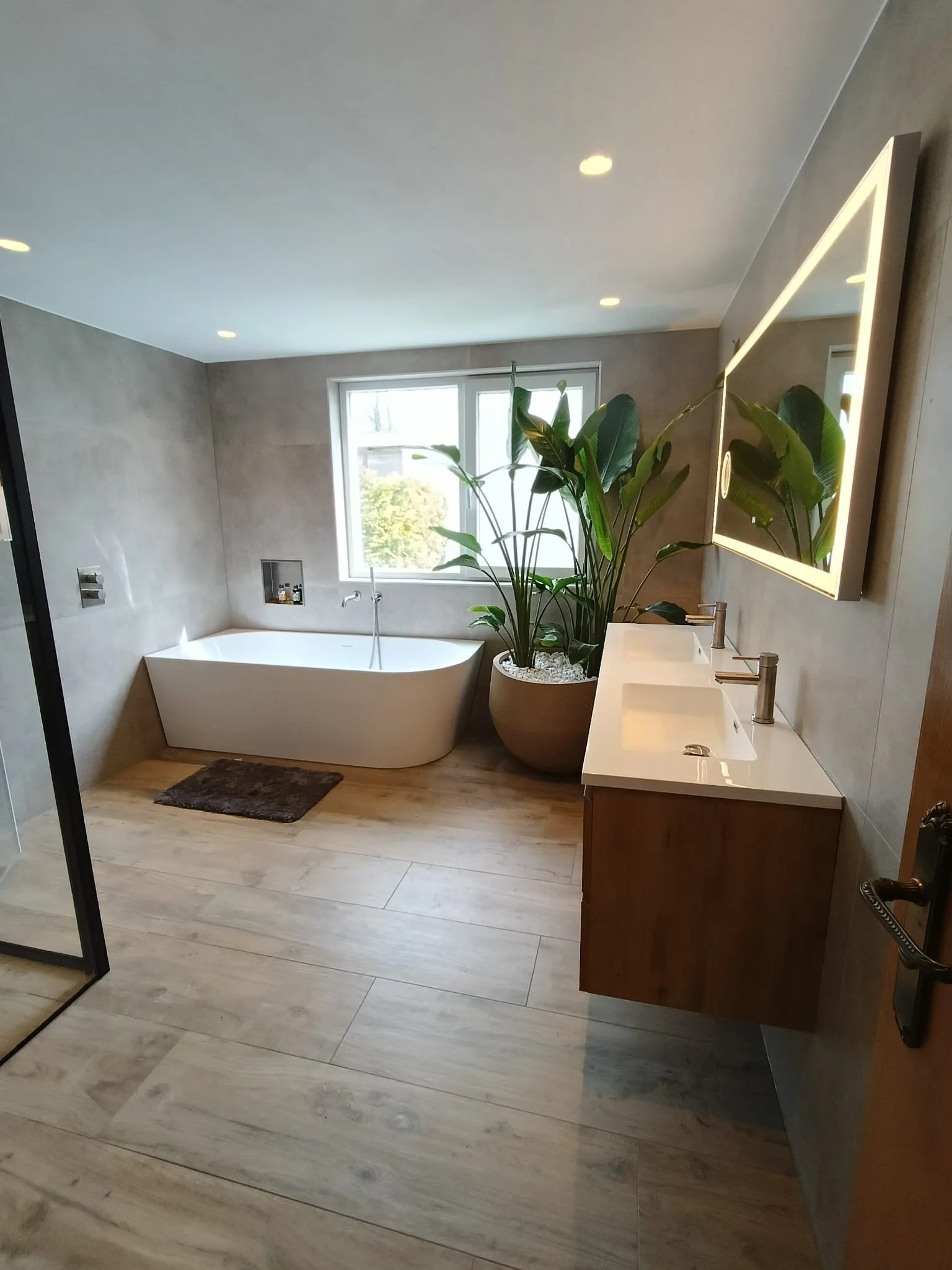 Modern badkamer met inloopdouche, ligbad, dubbele wastafel, grote spiegels met verlichting, grote ramen, planten en houten vloer.