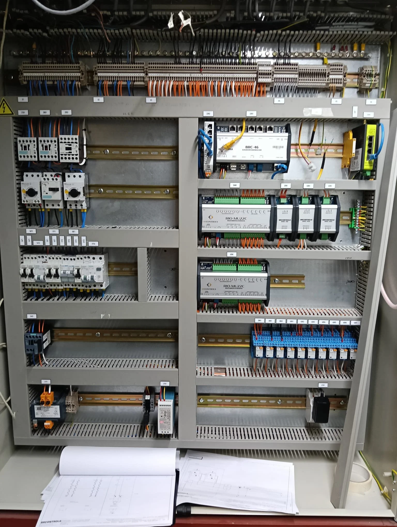 Binnenkant van een besturingskast met elektrische componenten en bedradings, documenten en papieren op een tafel.