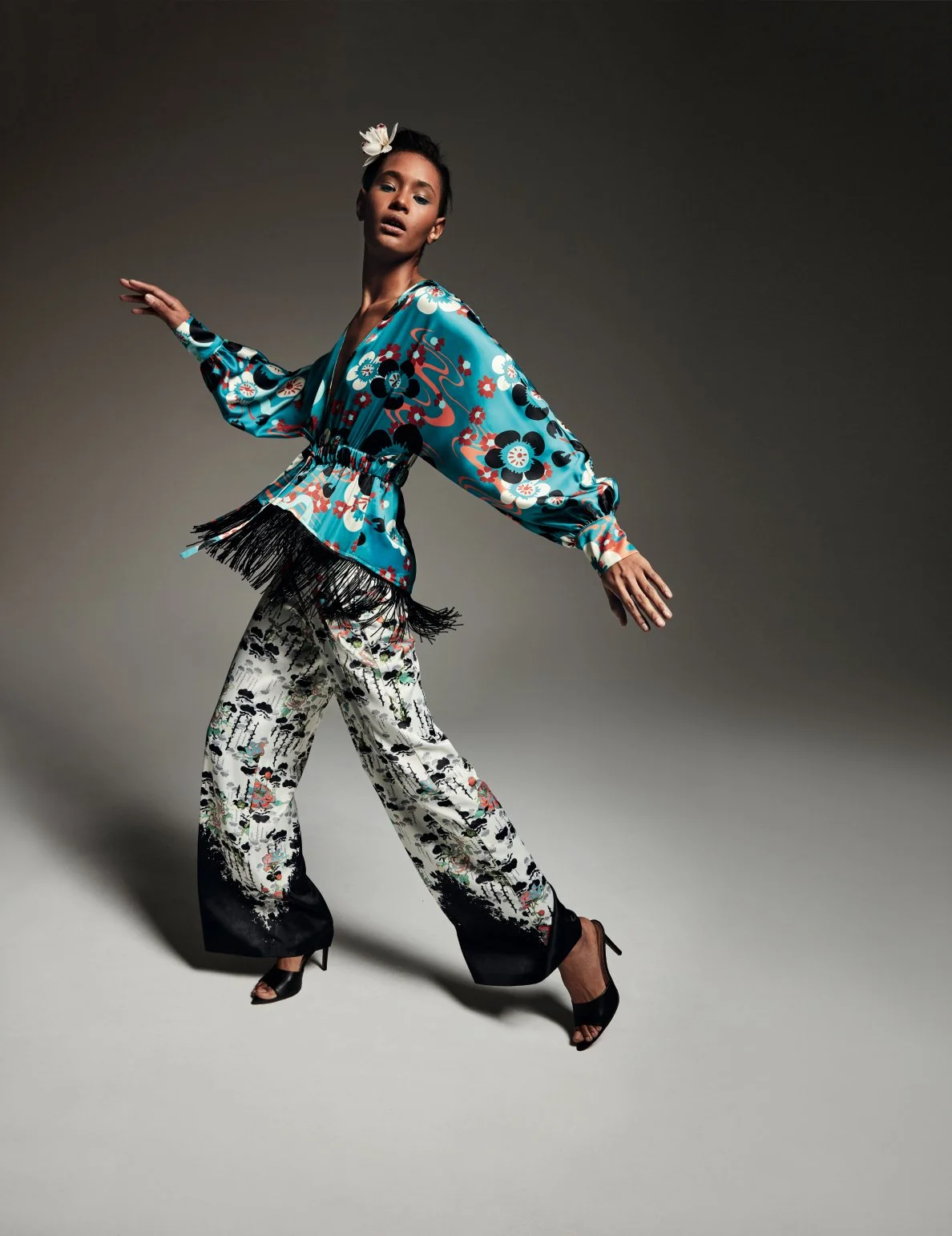 A woman striking a pose wearing a vibrant, floral patterned outfit with a blue kimono-style top, black and white wide-leg pants with floral details, black high heels, and a white flower in her dark hair, against a gray studio background.
