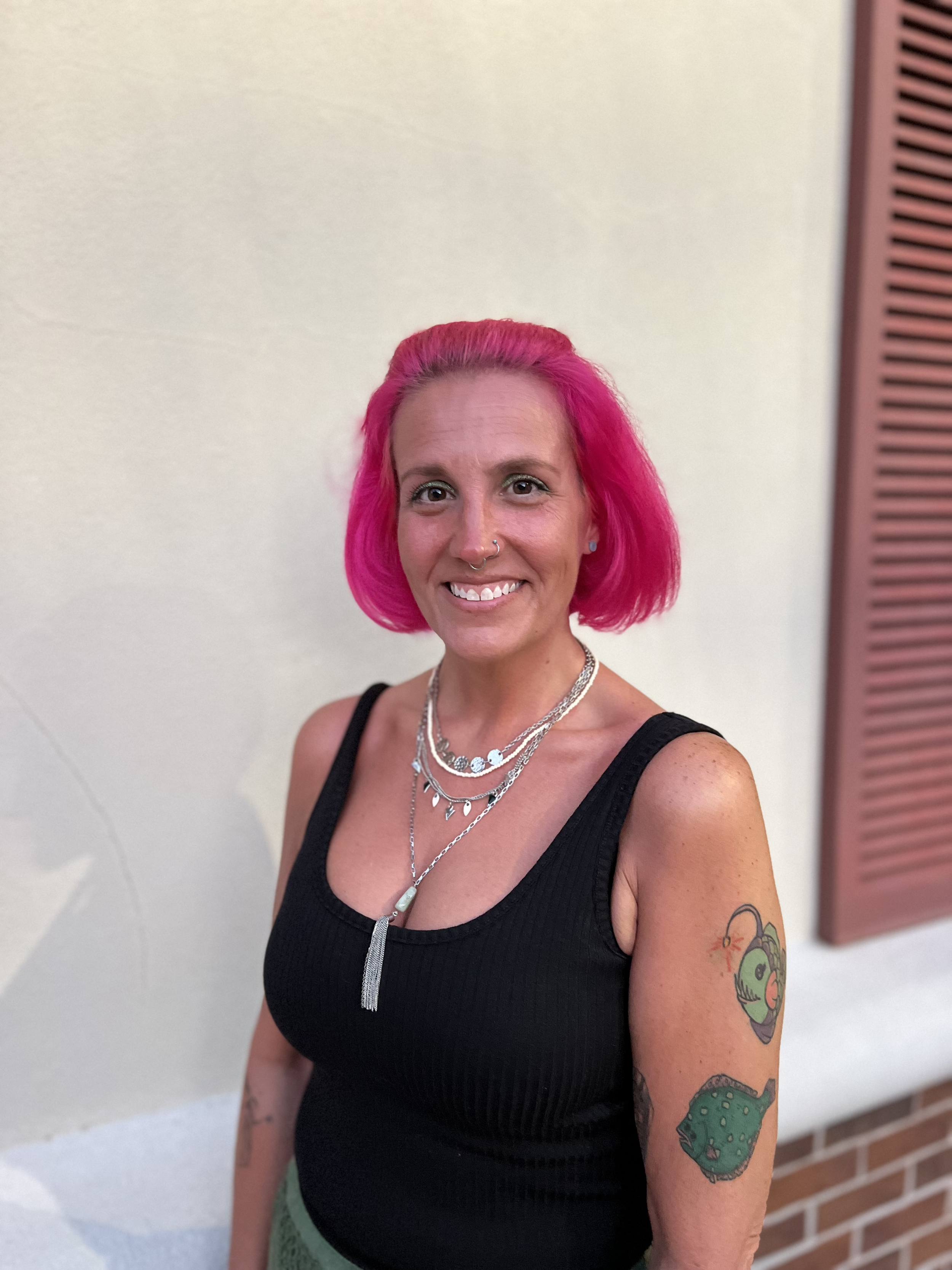 Woman with pink hair standing on a beach at sunset, smiling, wearing a white sleeveless top, red skirt, and a necklace with a tree-shaped pendant.