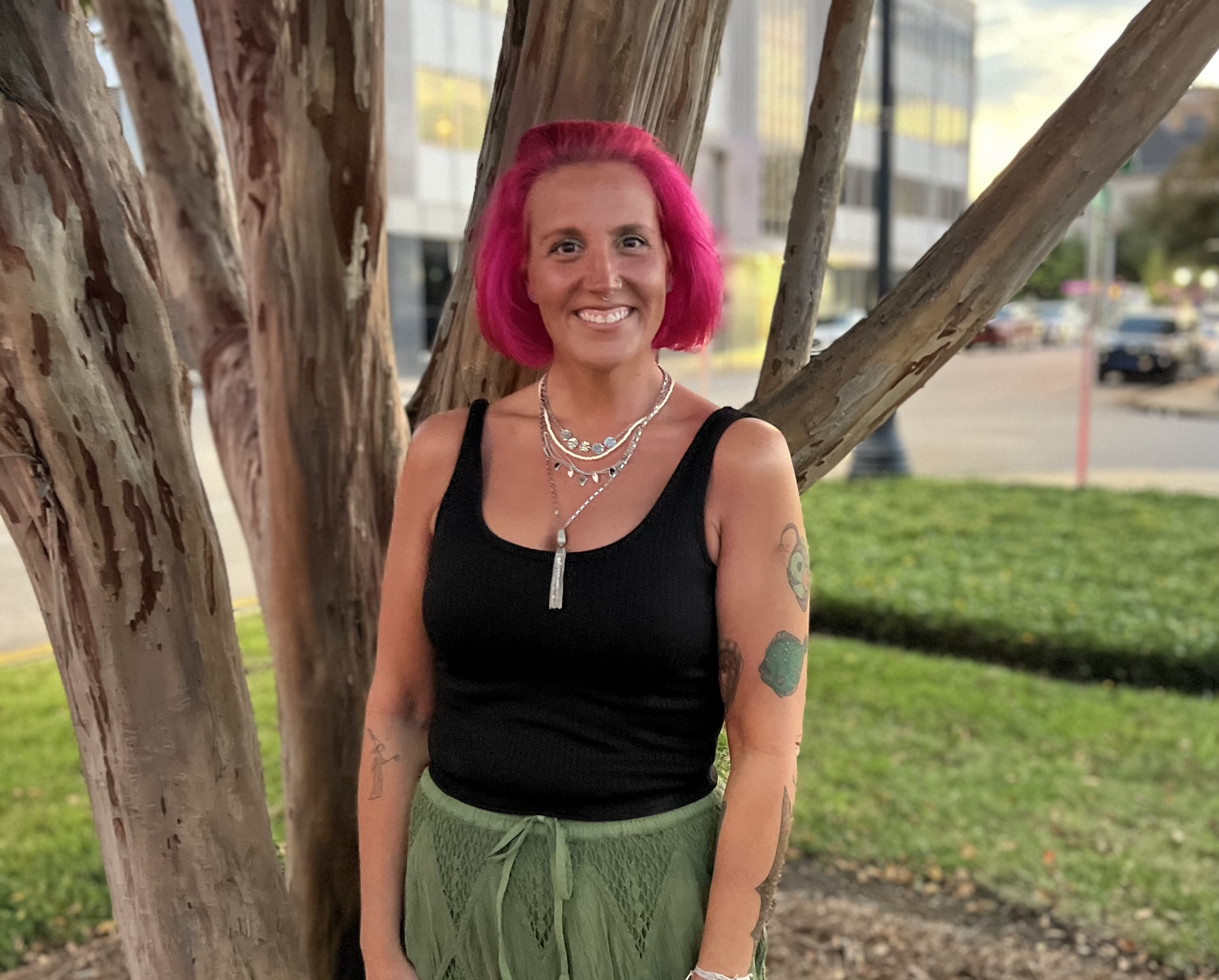 Woman with pink hair smiling on a beach at sunset, wearing a white sleeveless top and red skirt, standing with hands on hips.