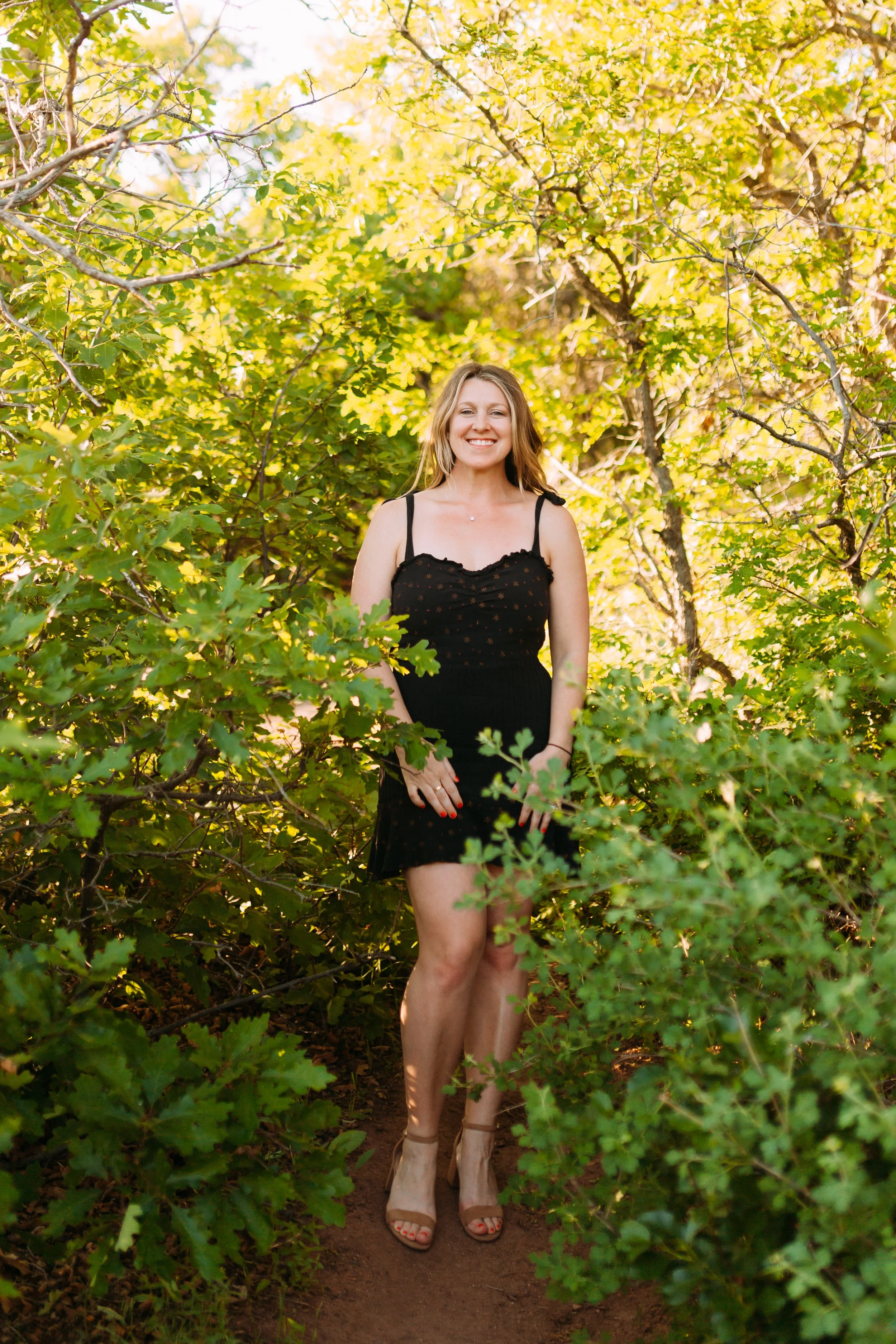 Woman standing on a dirt trail surrounded by green bushes and trees, smiling at the camera, wearing a black dress and beige heels.