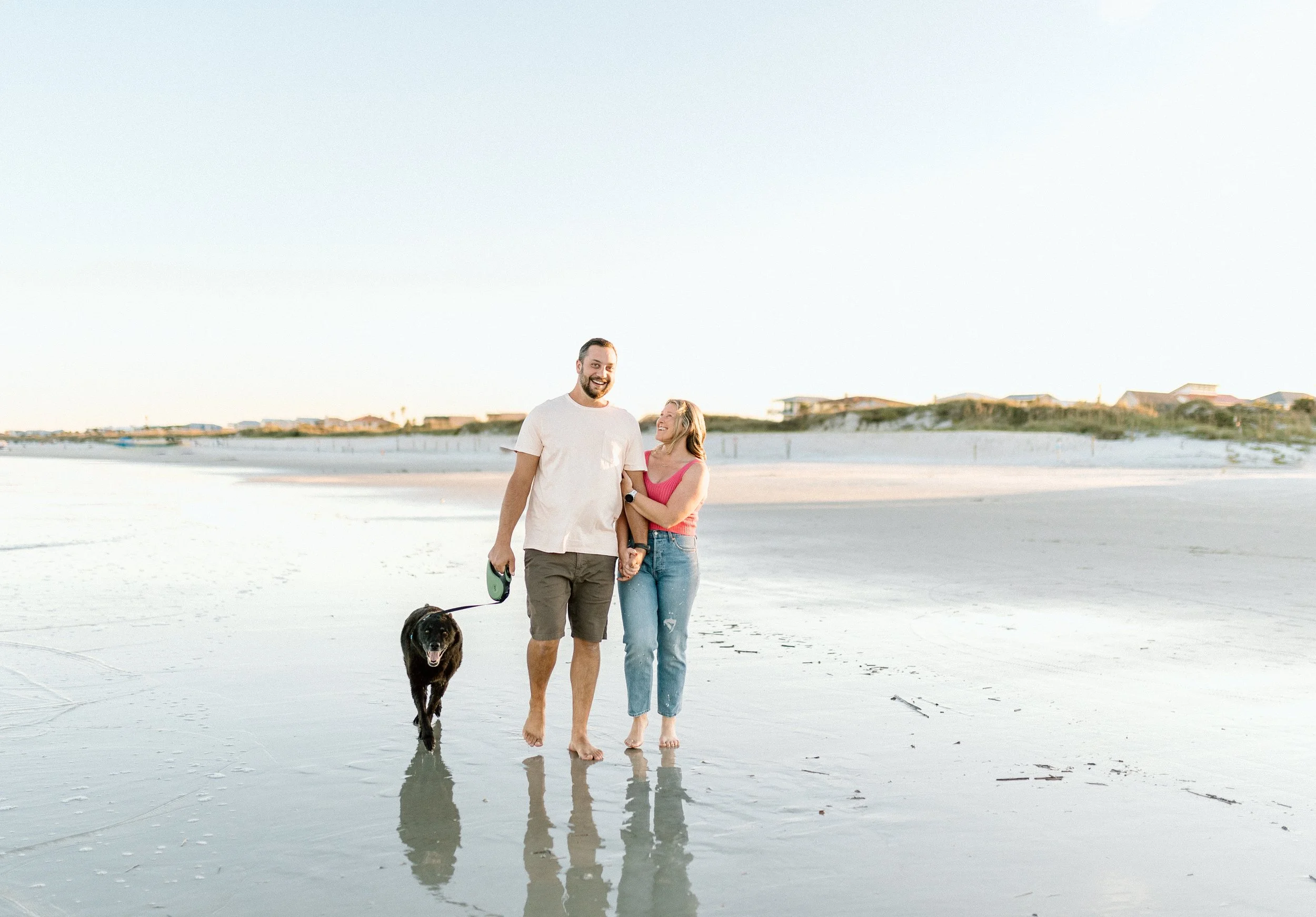 A smiling man and woman walking barefoot on the beach with their black dog during sunset, holding hands and enjoying the seaside.
